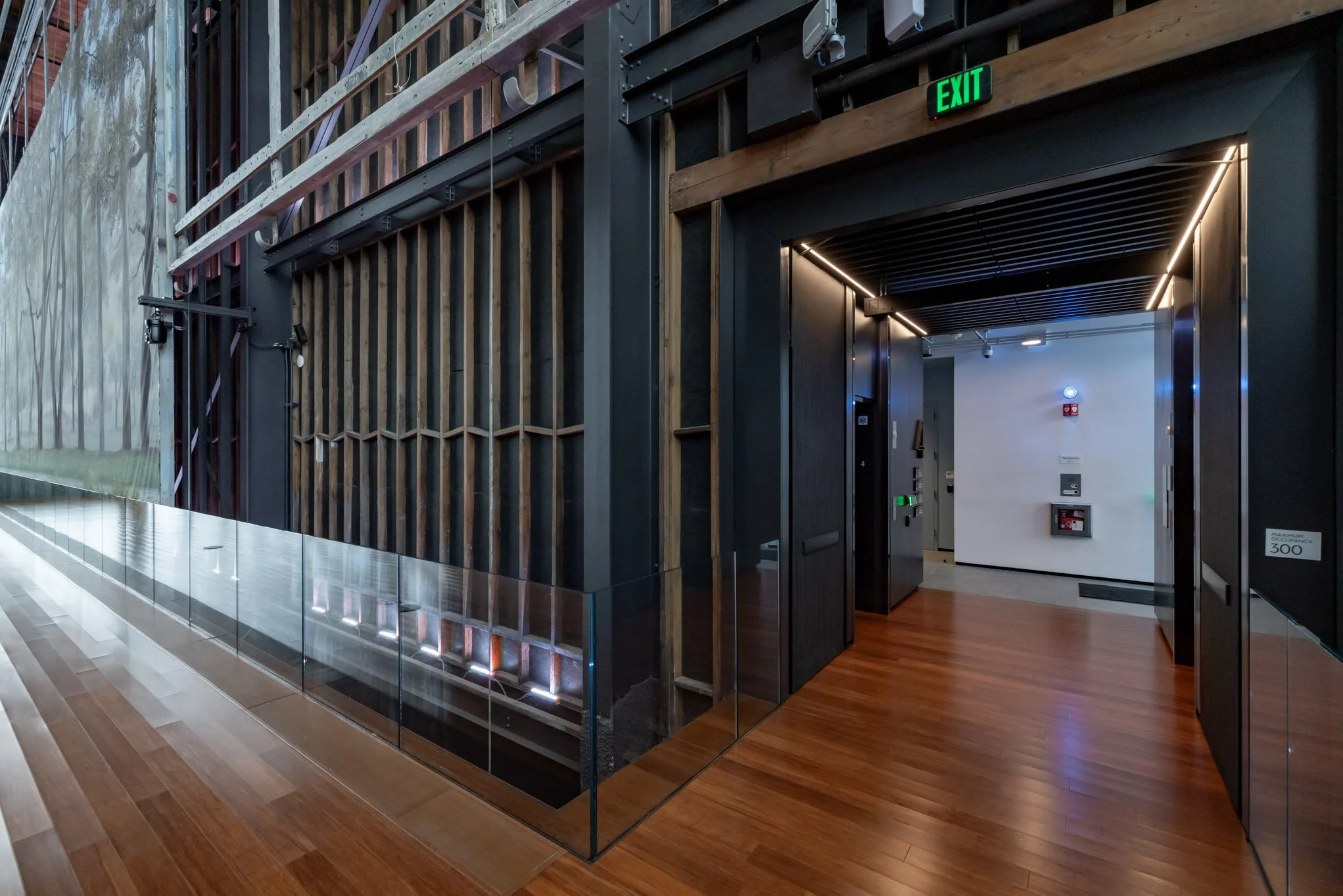 Modern interior space with wooden flooring and black structural elements. An open corridor leads to an elevator with a green emergency exit sign above it. There's a glass panel on the left side, and the walls are decorated with vertical wood slats an