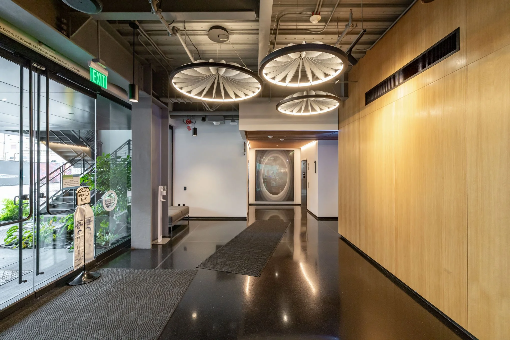Modern hotel lobby with black polished floor, wooden wall, ceiling lights, glass entrance door, bench, plants, and elevator in the background.