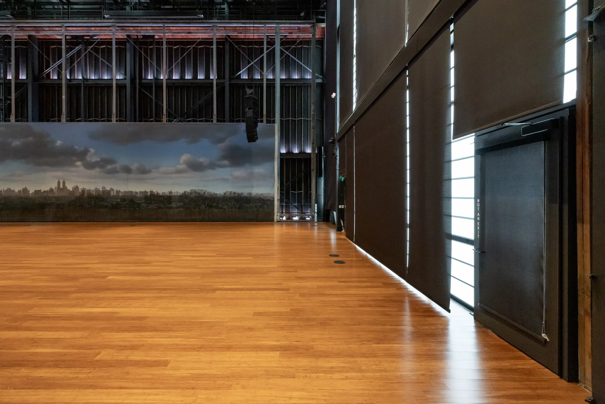 An empty indoor room with a wooden floor, black walls with vertical light strips, and a large window with blinds, possibly a stage or event space.
