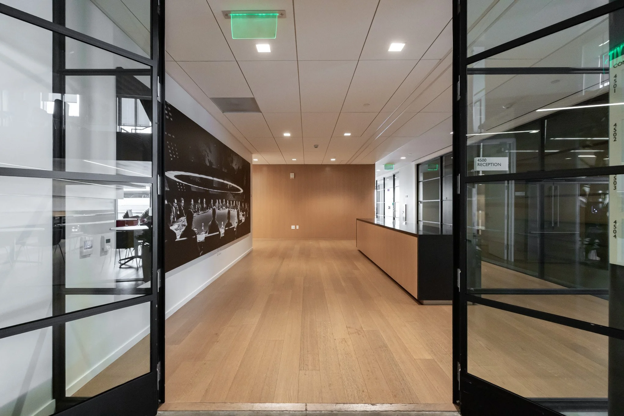 Empty office reception area with wooden floors, glass walls, and a black and white photo on the wall. Glass doors with green exit signs, and a reception desk to the right.