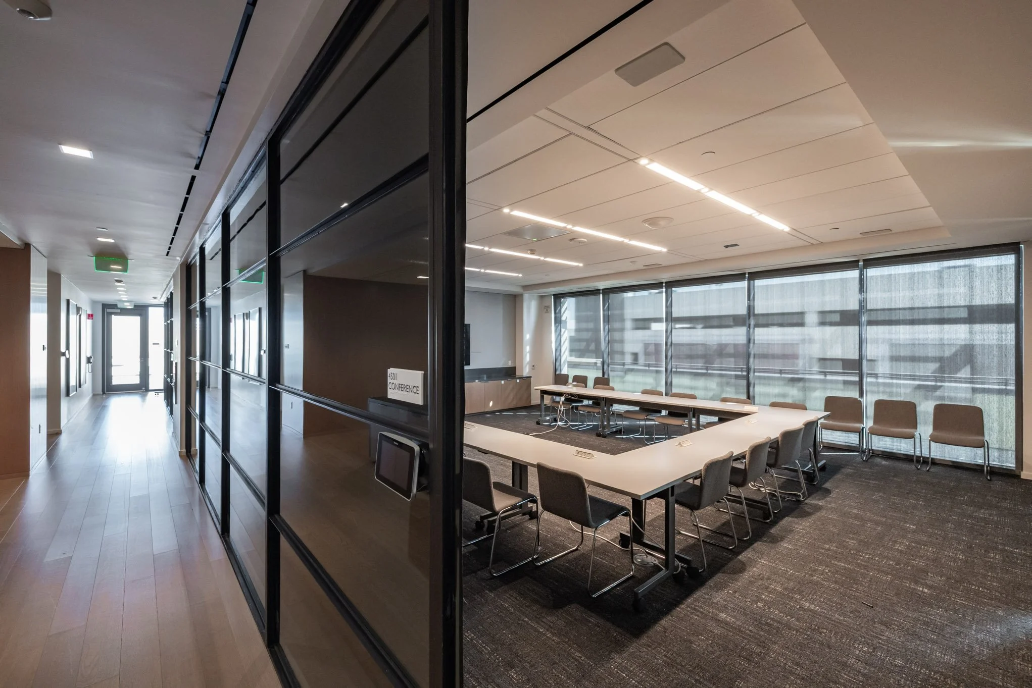 Empty conference room with large windows, U-shaped table and chairs, and modern ceiling lighting in a corporate office building.
