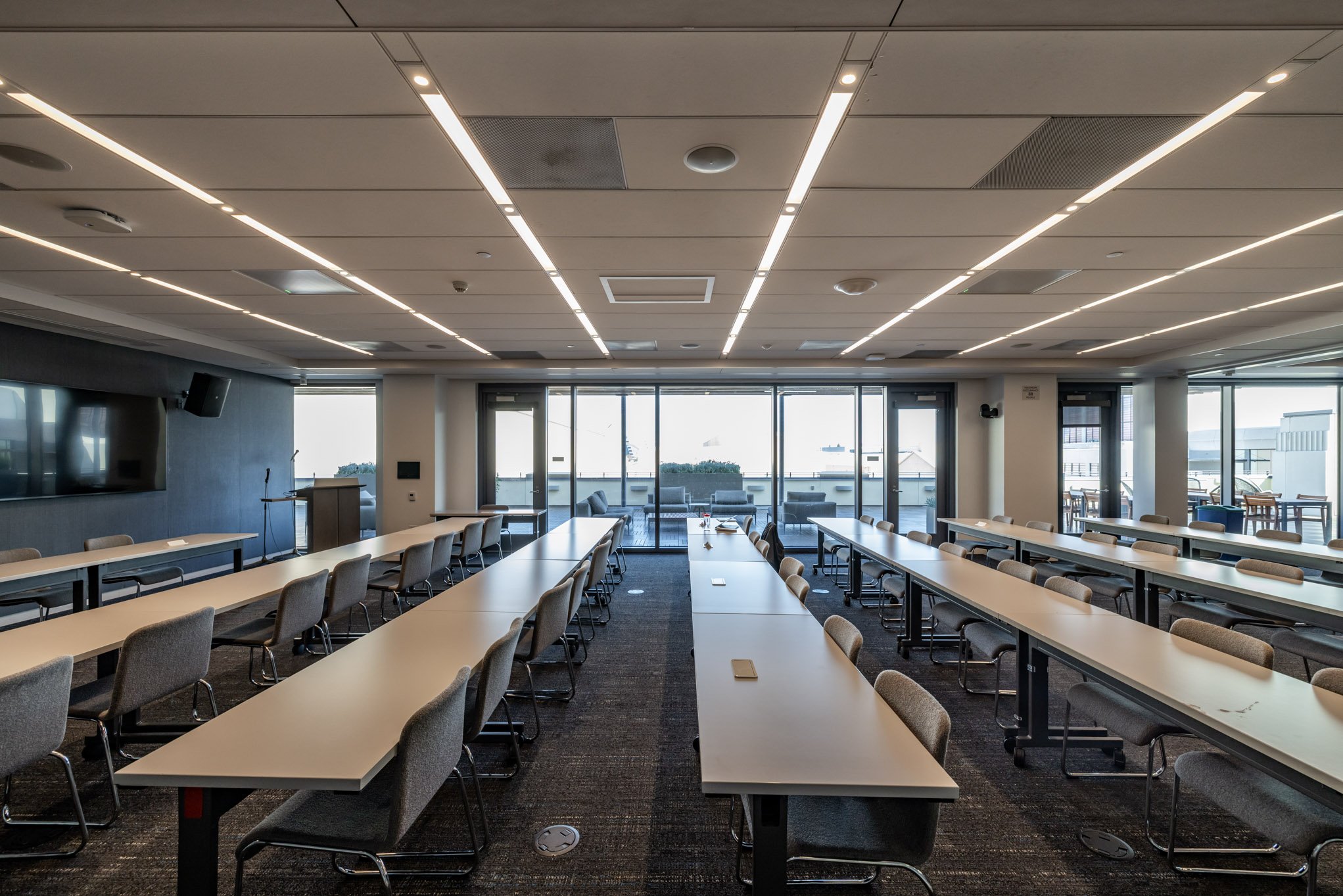 Empty conference room with rows of tables and chairs, large windows overlooking a balcony with outdoor seating, ceiling with recessed lighting.