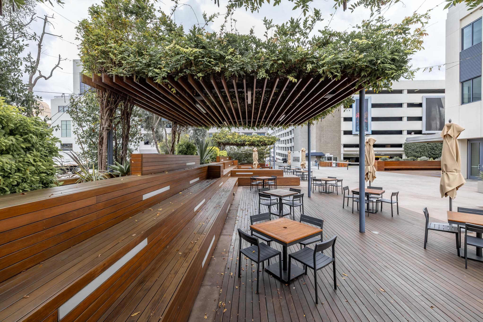 Empty outdoor patio with wooden flooring, tables, and chairs, surrounded by bushes and trees, with umbrellas and modern buildings in the background.