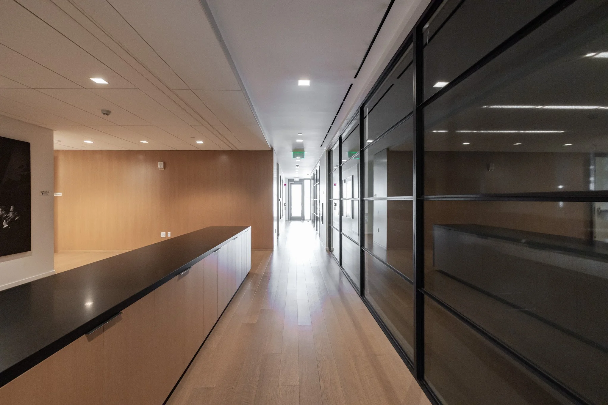 A modern indoor hallway with wooden floors, glass walls on the right, and a wooden wall on the left, leading to a light-filled exit door at the end.