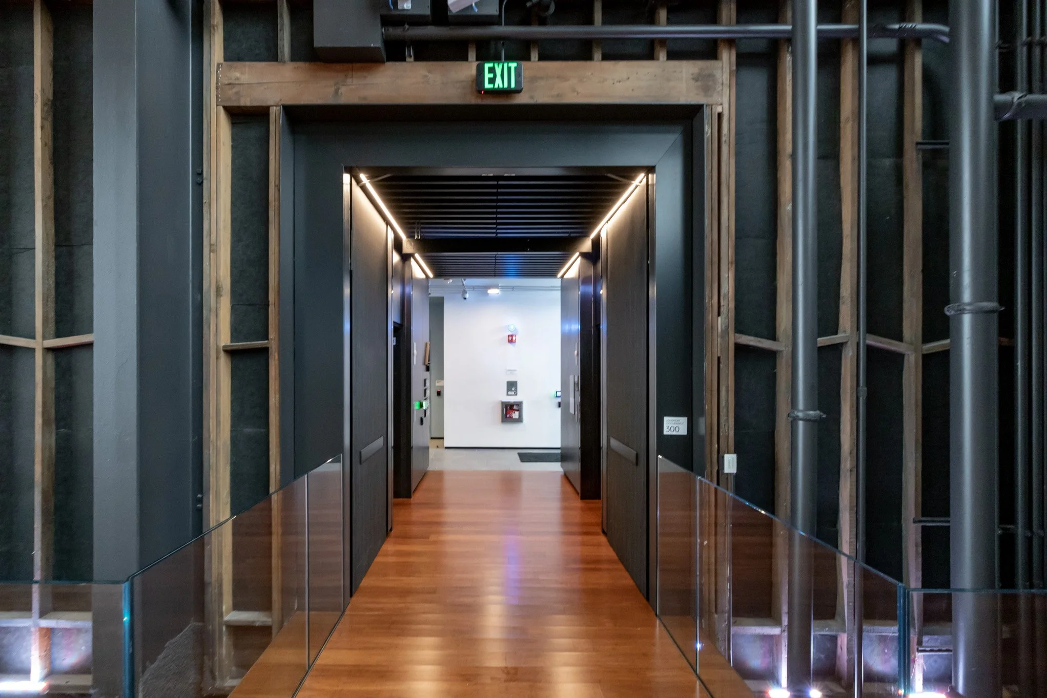Indoor hallway with wooden flooring, black walls, and lighting leading to an exit door marked with a green "EXIT" sign.