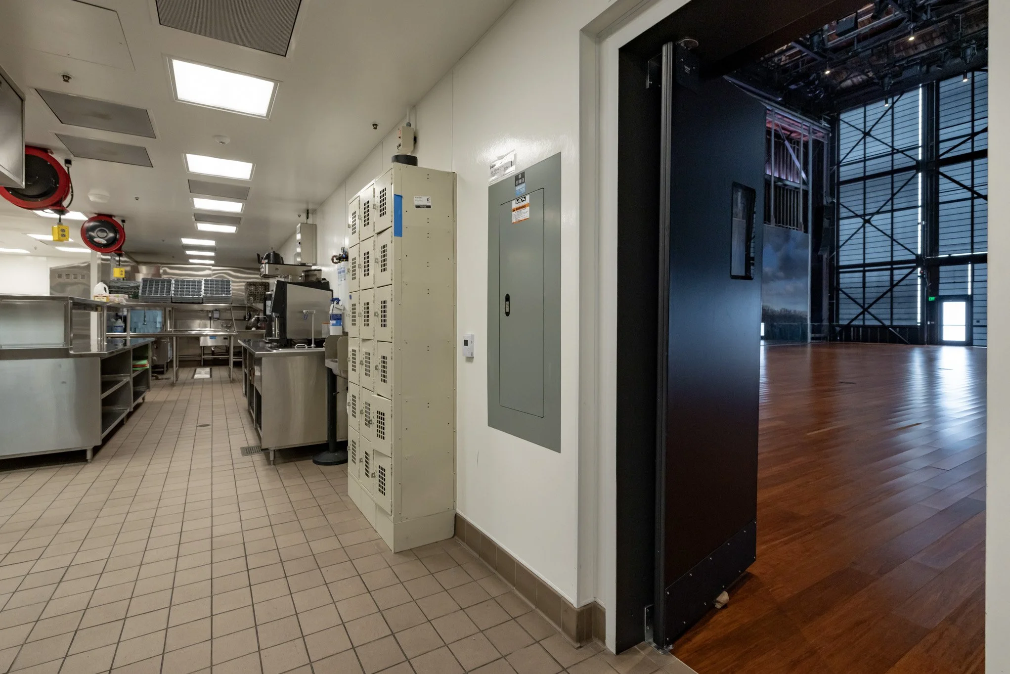 Interior view of a commercial kitchen with stainless steel appliances and storage; doorway leading to a stage area with wooden flooring and large windows.