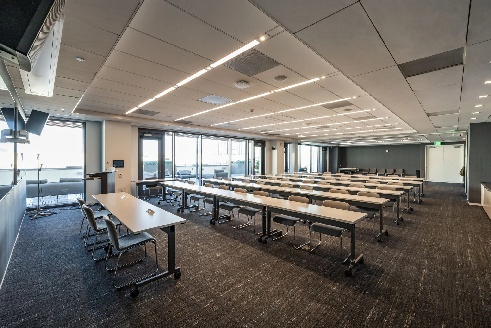 An empty conference room with rows of tables and chairs, large windows with outdoor views, and ceiling lights.