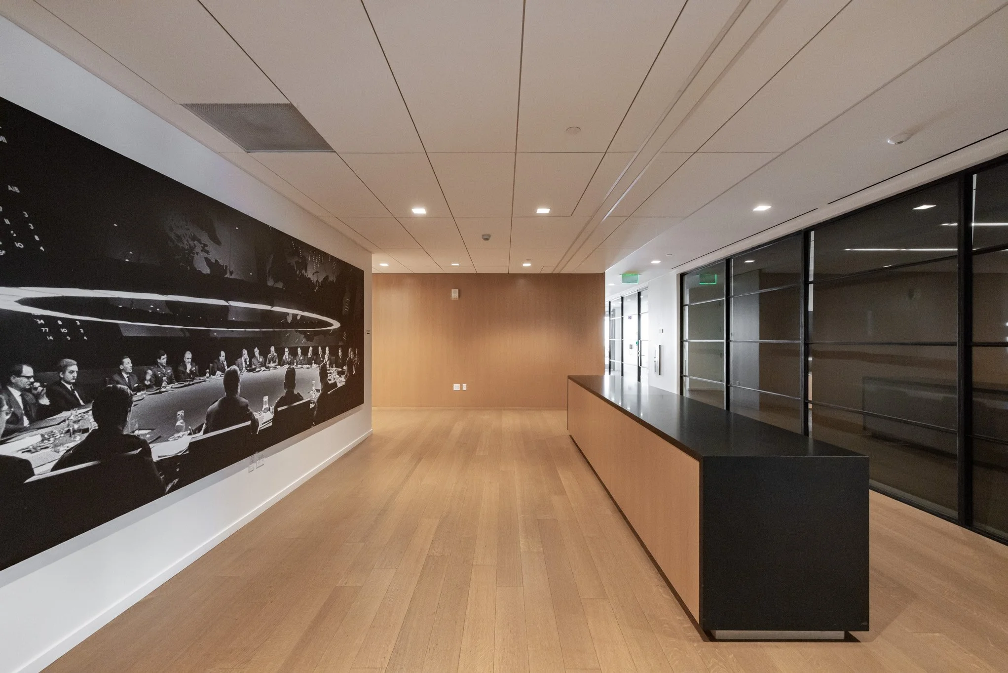Empty hallway in modern office building with a large black and white mural of a business meeting on the left wall, wooden accents, and glass walls on the right.