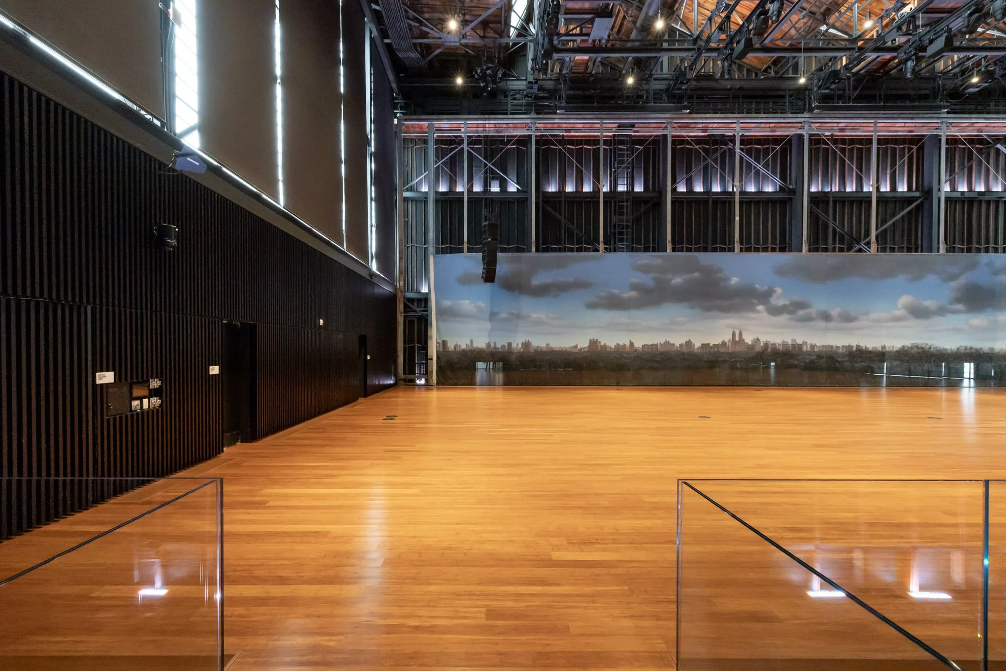 Empty indoor sports or event hall with wooden floor, black wall panels, and a large cityscape mural on the back wall. Metal scaffolding structure is visible near the ceiling.