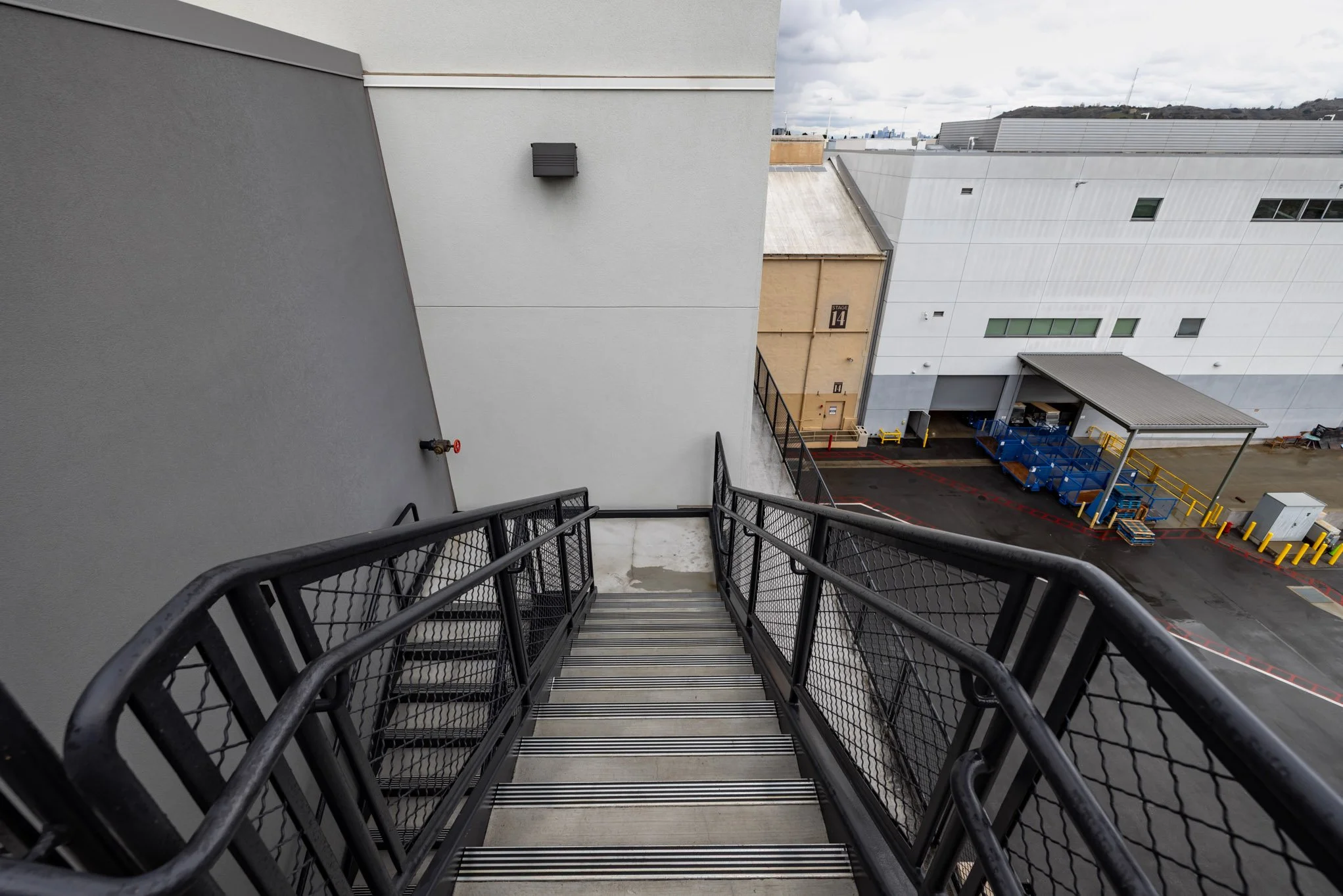 View from top of an outdoor metal staircase looking down at a loading dock area with blue carts, yellow safety barriers, and warehouse buildings in the background.