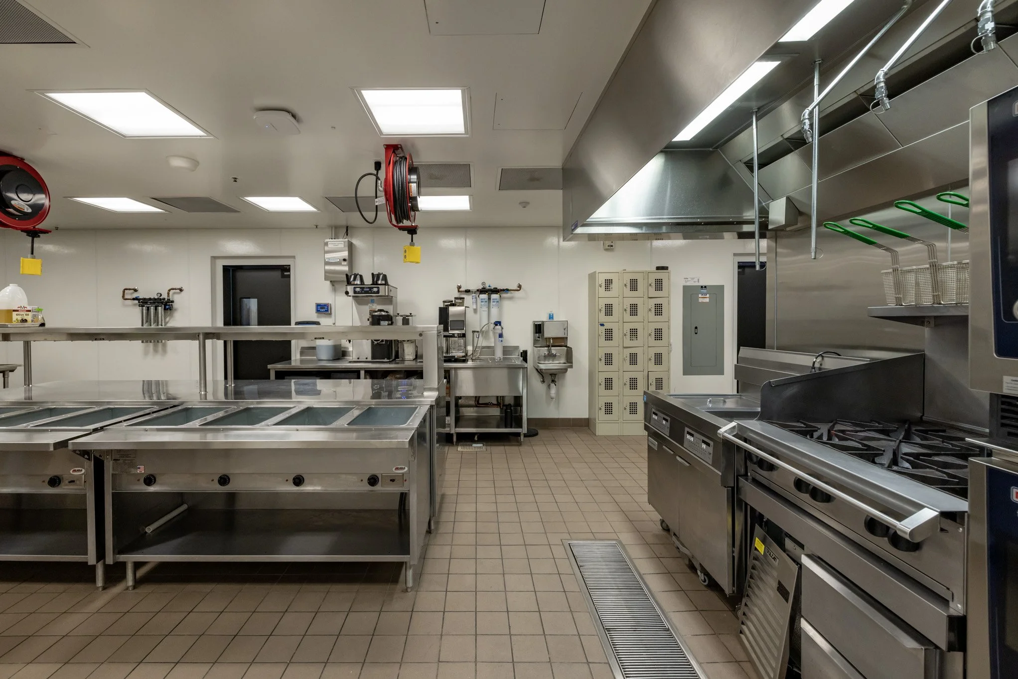 Empty commercial kitchen with stainless steel appliances, counters, and lockers. Fireplace hoses and kitchen tools are also visible.