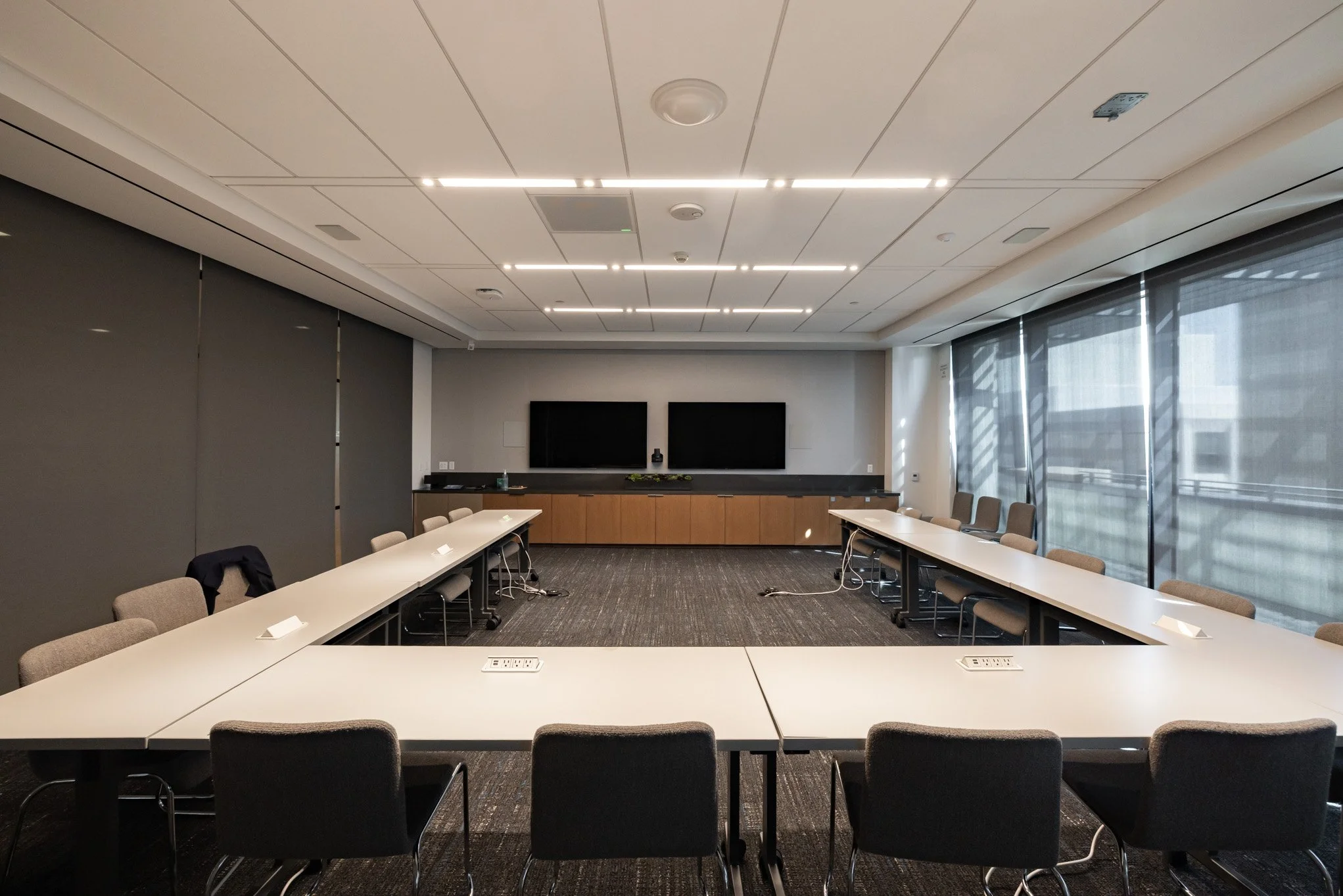 Empty modern conference room with U-shaped tables, gray chairs, large windows with blinds, and two large black screens at the front.