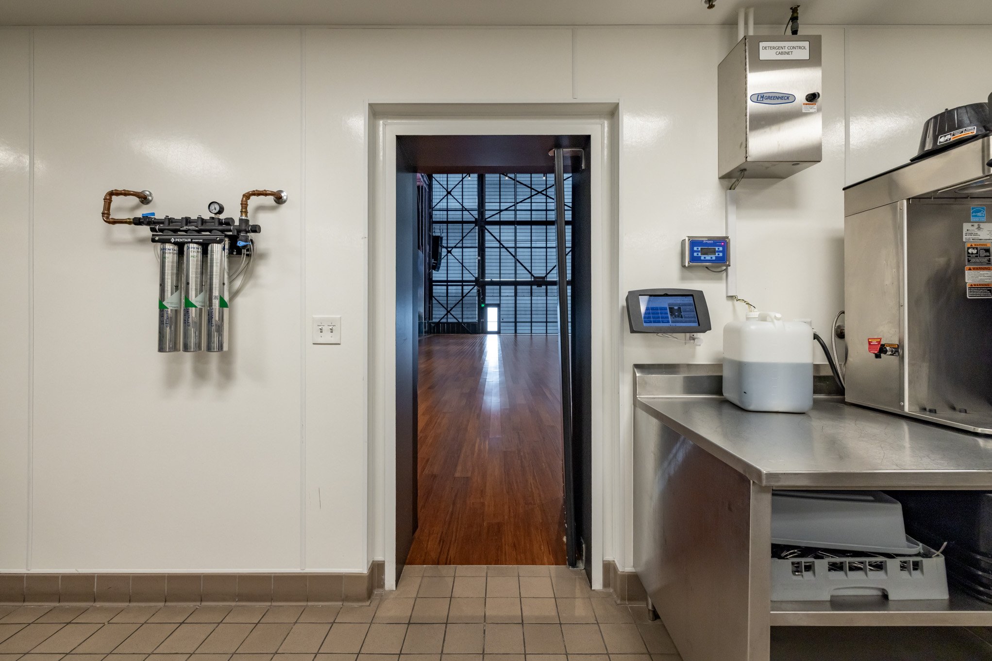 Industrial kitchen with filtration system on the wall, touchscreen controls, and a white container on a stainless steel countertop. There's a door leading to a large open space with wooden flooring and scaffolding outside.