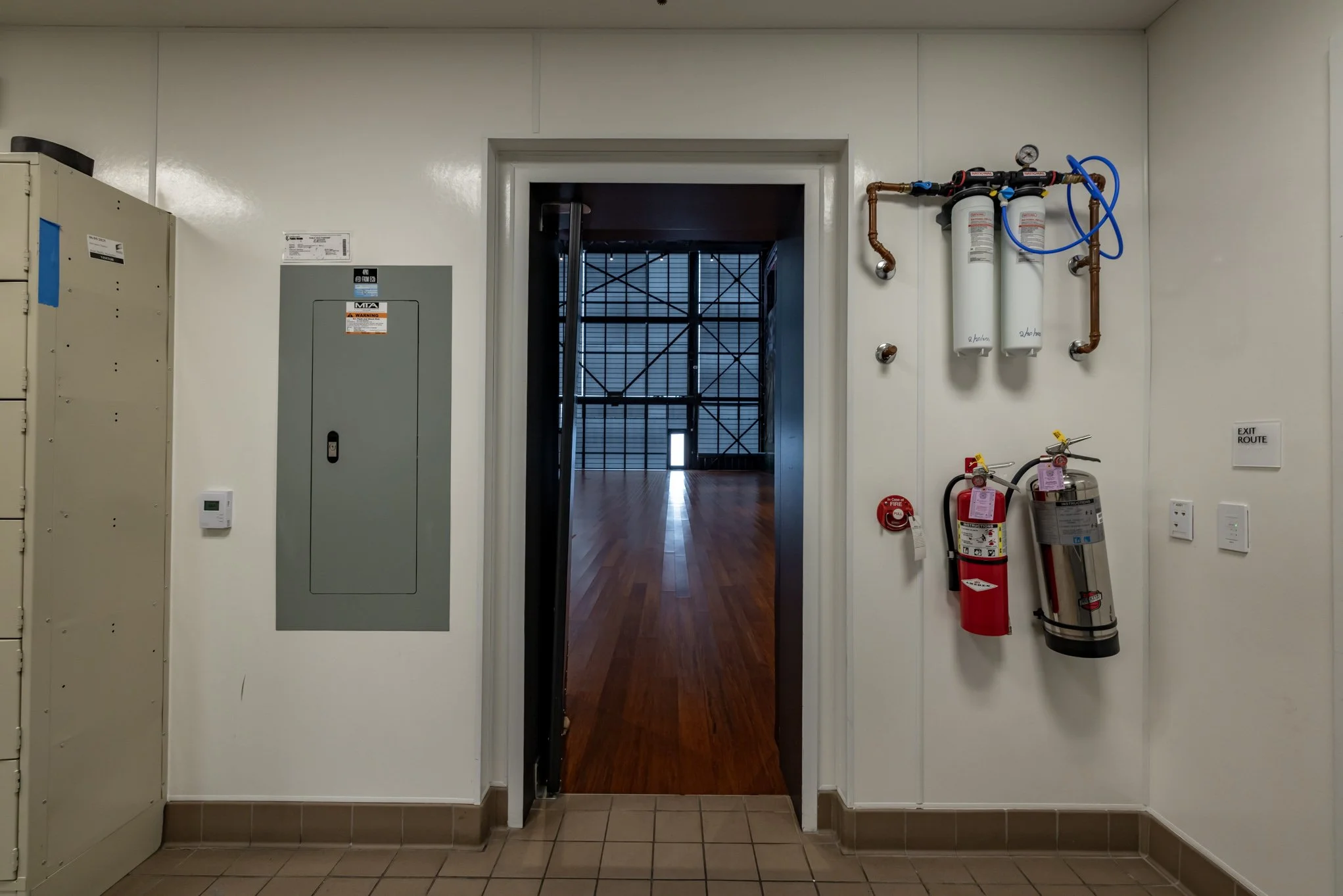Interior view of a building hallway with a door leading to a room with wooden floors and scaffolding outside. Fire safety equipment including fire extinguishers and fire alarms are mounted on the white walls, and an electrical panel is also visible.
