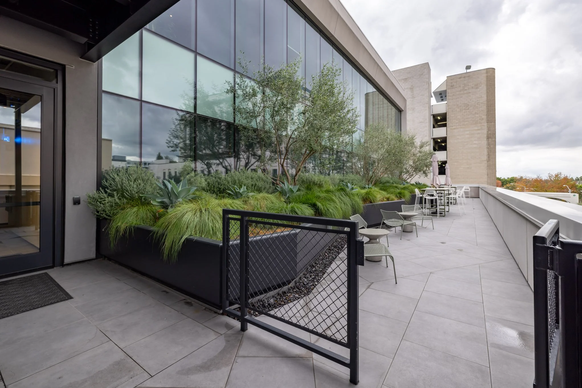 Outdoor balcony with modern seating and planters, large glass windows, and a view of trees and city buildings.
