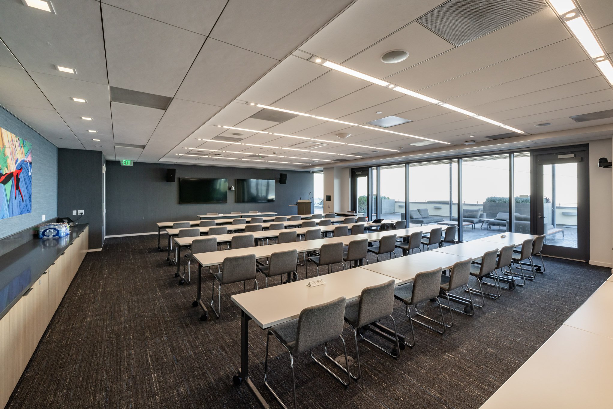 A modern conference room with multiple blank tables and gray chairs facing a wall with two large flat-screen TVs, large windows on one side with outdoor seating, and a ceiling with recessed lighting.