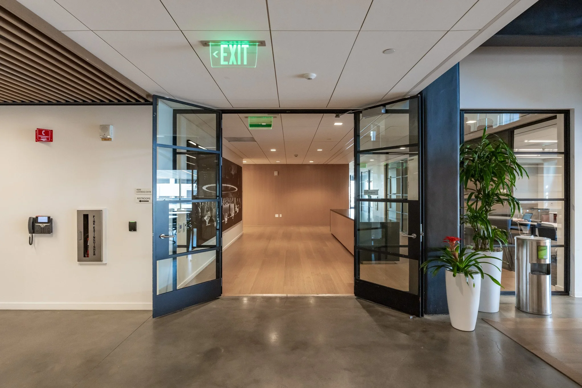 Open doors to a modern conference center or office space with plants, a fire extinguisher, and exit signs.