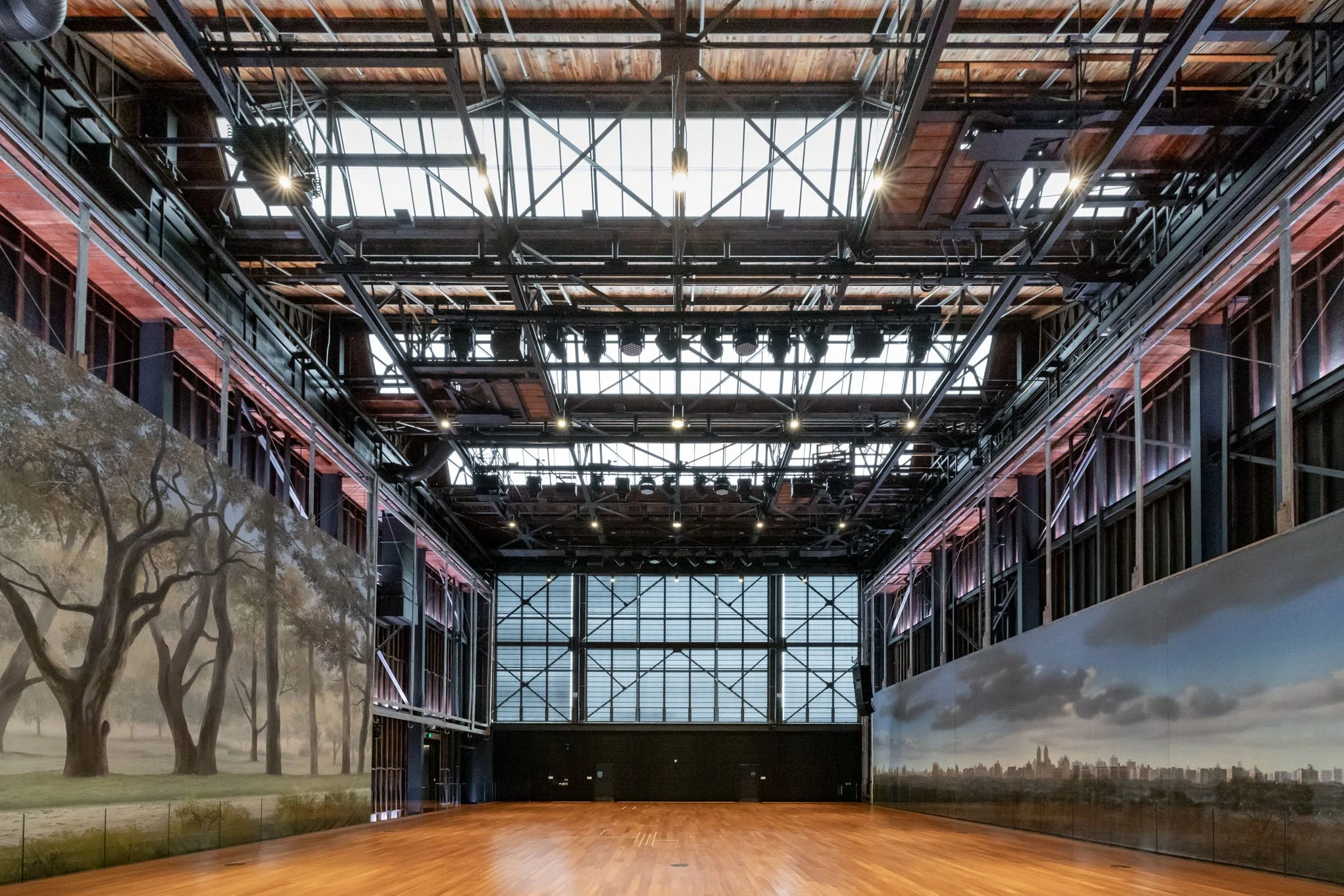 An empty indoor event space with wooden flooring, mural paintings of trees and cityscapes on the walls, and a metal and wooden ceiling with skylights and stage lighting.