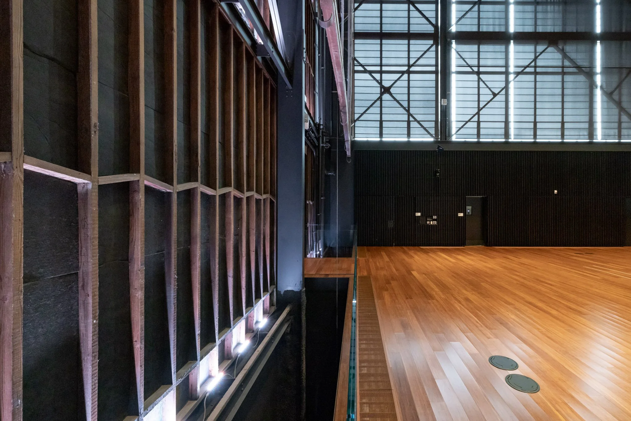 Empty indoor gymnasium with wooden flooring, black walls, large windows with metal framing, and ceiling lights