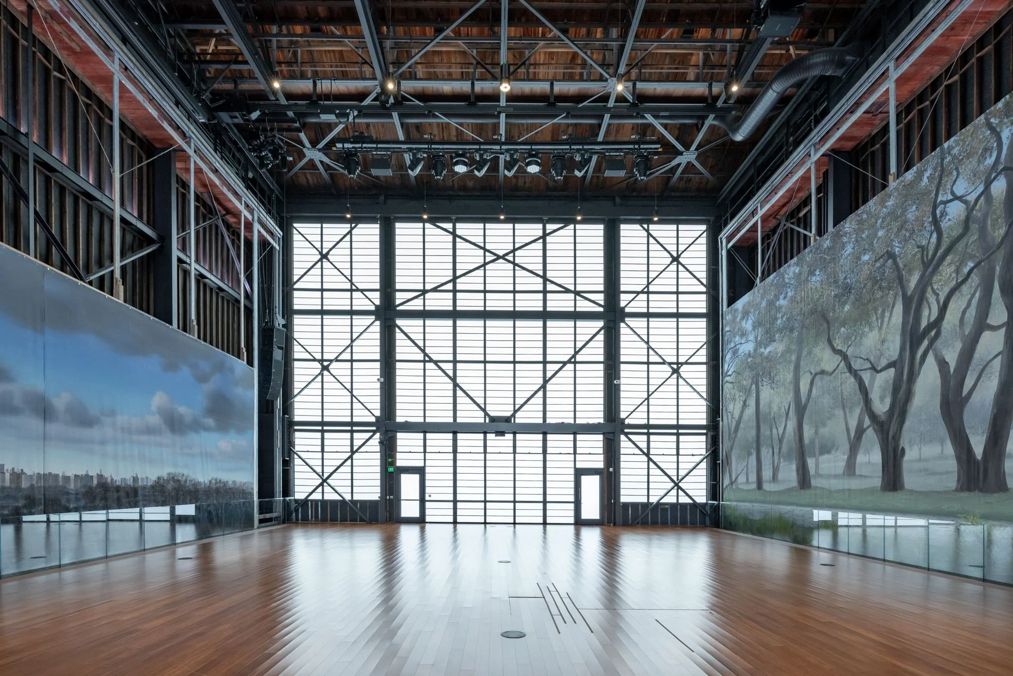 Empty indoor gallery with large glass window doors, wooden flooring, and painted murals of trees and landscape on the walls.