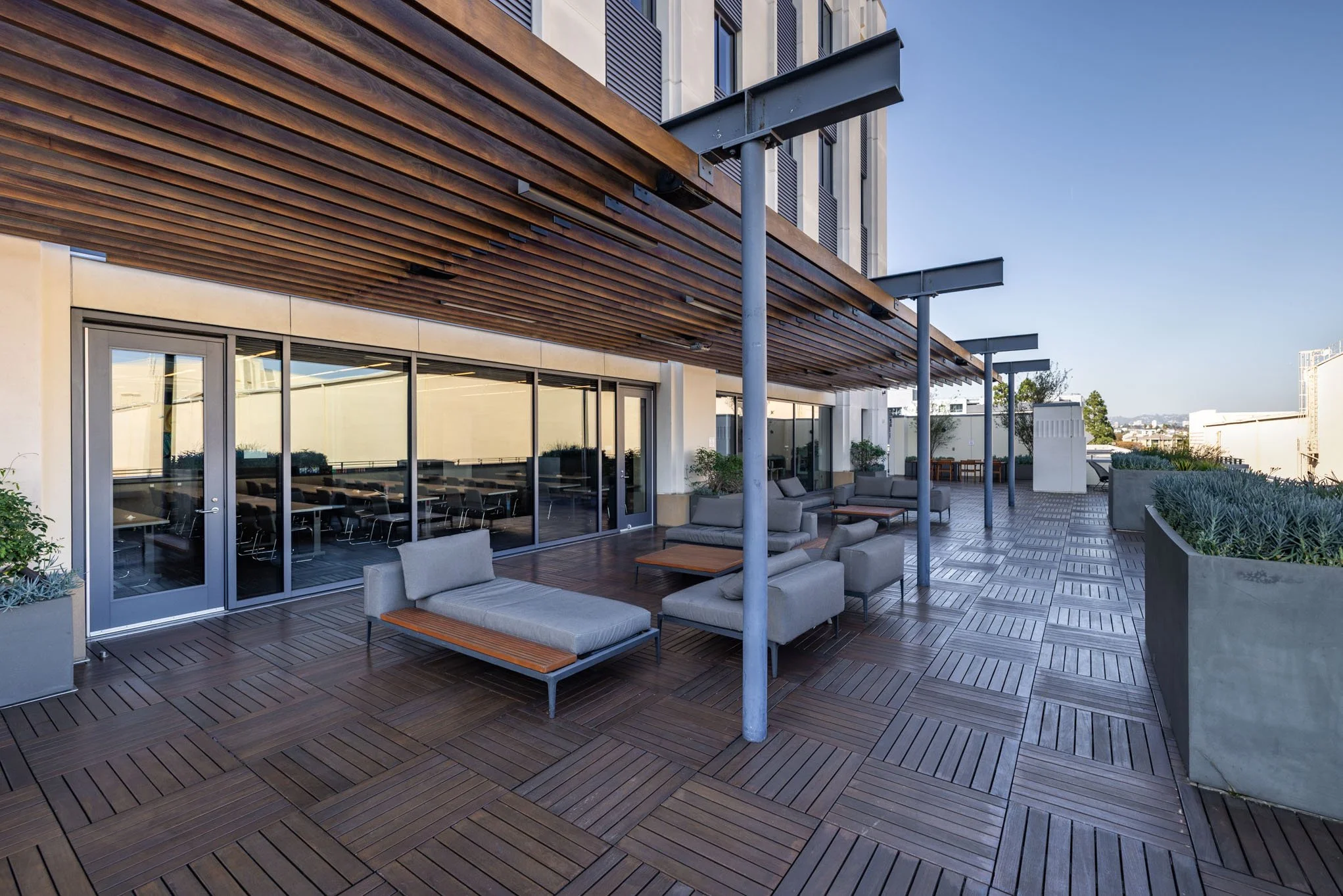 Outdoor balcony or rooftop terrace of a modern building with wooden flooring, lounging furniture, and large glass doors leading inside.