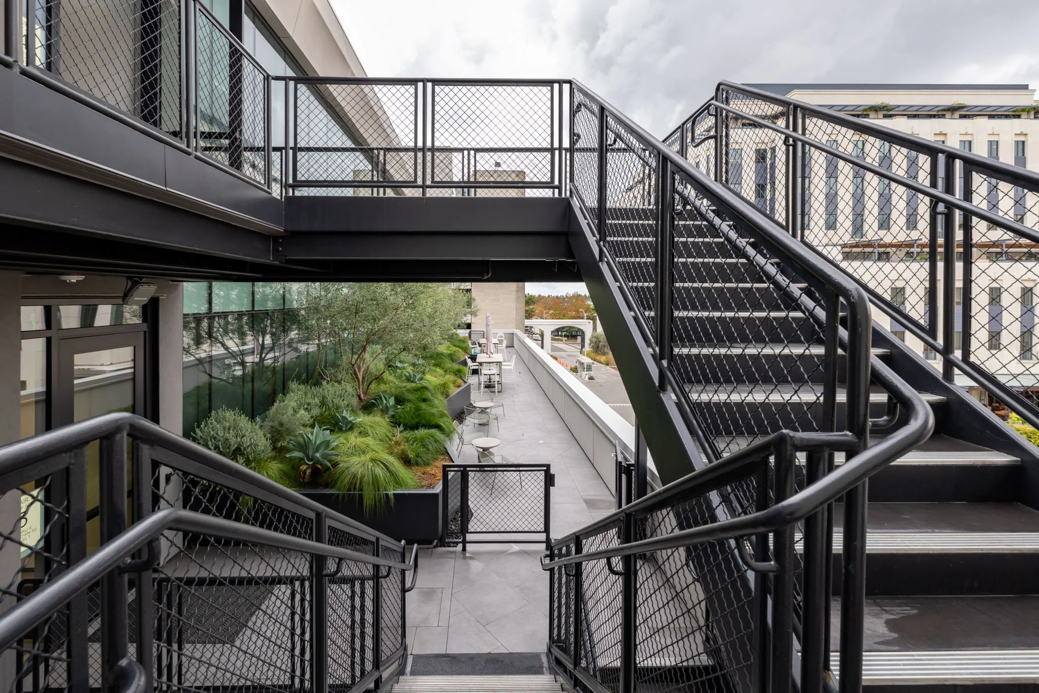 Outdoor balcony area with stairs and lush green plants along the railing, in an urban setting with modern buildings.