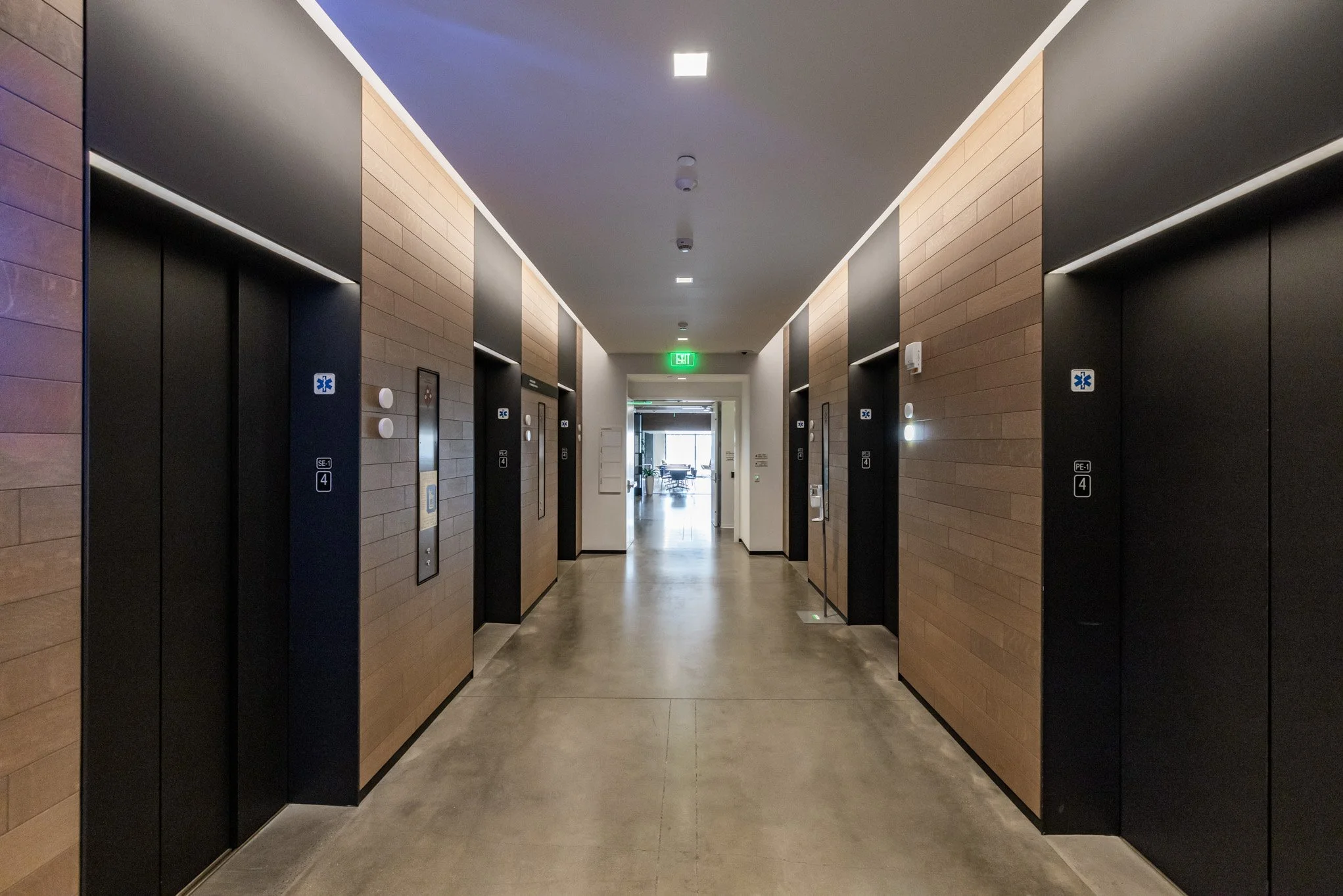 Hallway with six elevator doors, wood panel walls, and a doorway at the end leading to a bright room with tables and chairs.