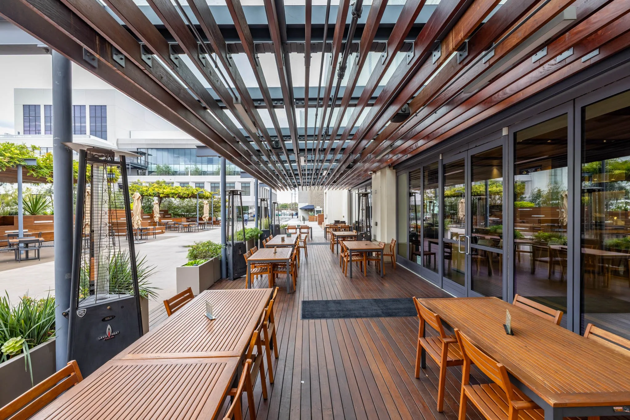 Empty outdoor restaurant patio with wooden tables and chairs, patio heaters, and modern building in the background.