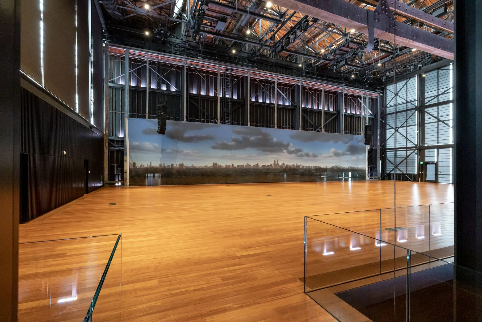An empty, large indoor space with a polished wooden floor, black walls, and a cityscape mural on the back wall. The ceiling has exposed metal framework and lighting fixtures.