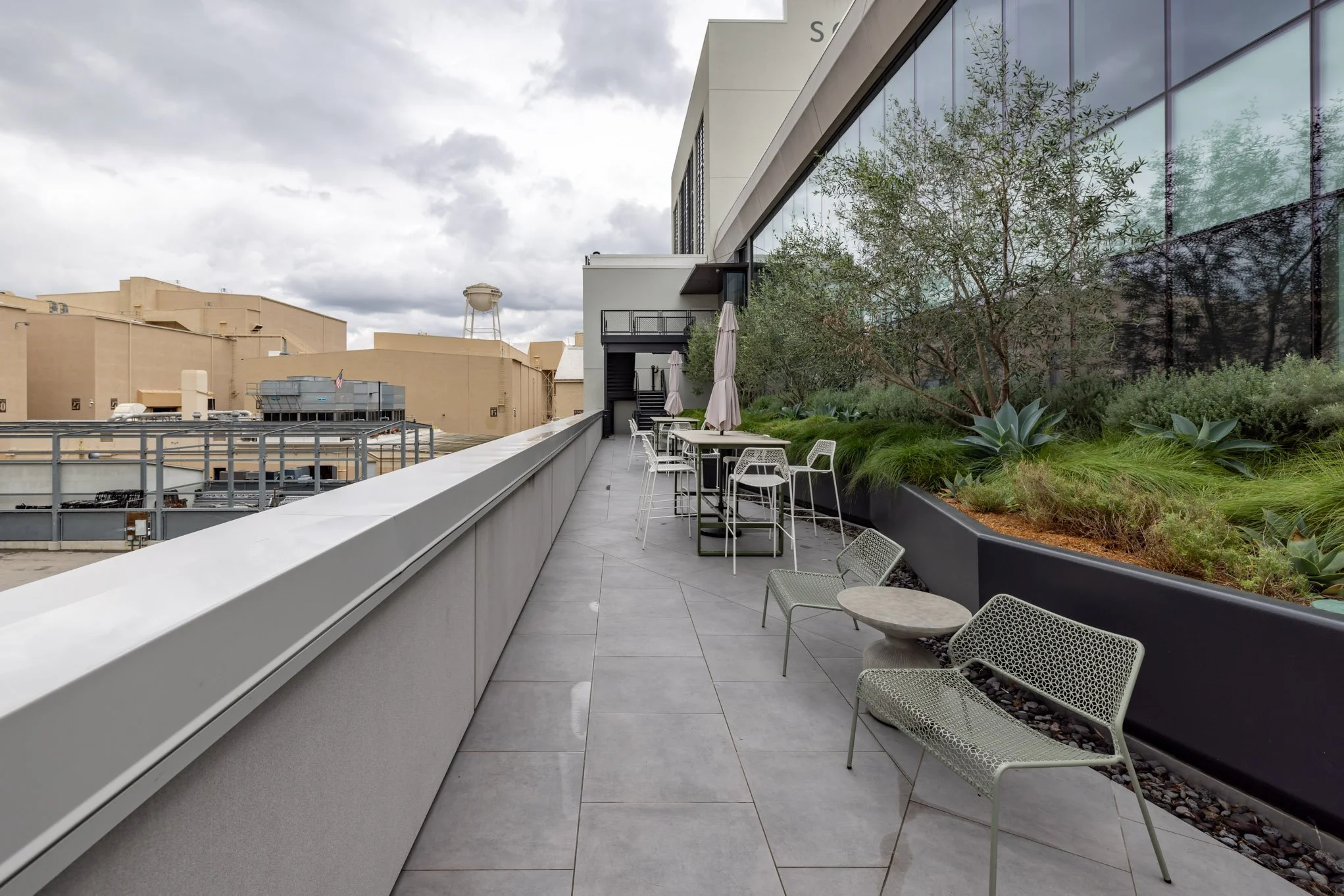 Empty outdoor terrace with chairs, tables, and potted plants, overlooking neighboring buildings and a cloudy sky.