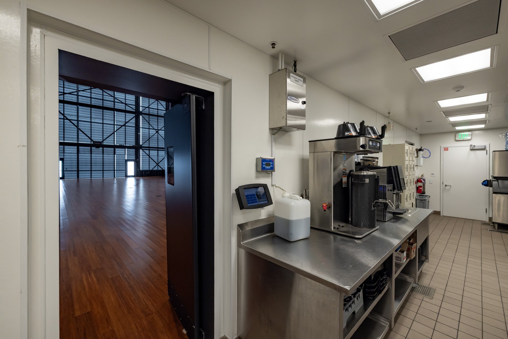 Commercial kitchen with coffee machines, a container, and kitchen appliances, adjacent to a door leading to a large, empty warehouse or studio space with wooden flooring and metal framework walls.
