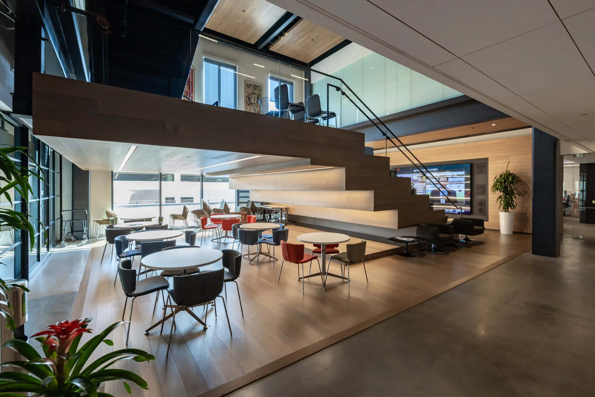 Modern lobby with wooden flooring, round tables, and chairs in gray, black, red, and green. A staircase with wooden steps and glass railing leads to an upper level with chairs and a window. Large windows let in natural light. A large television scree