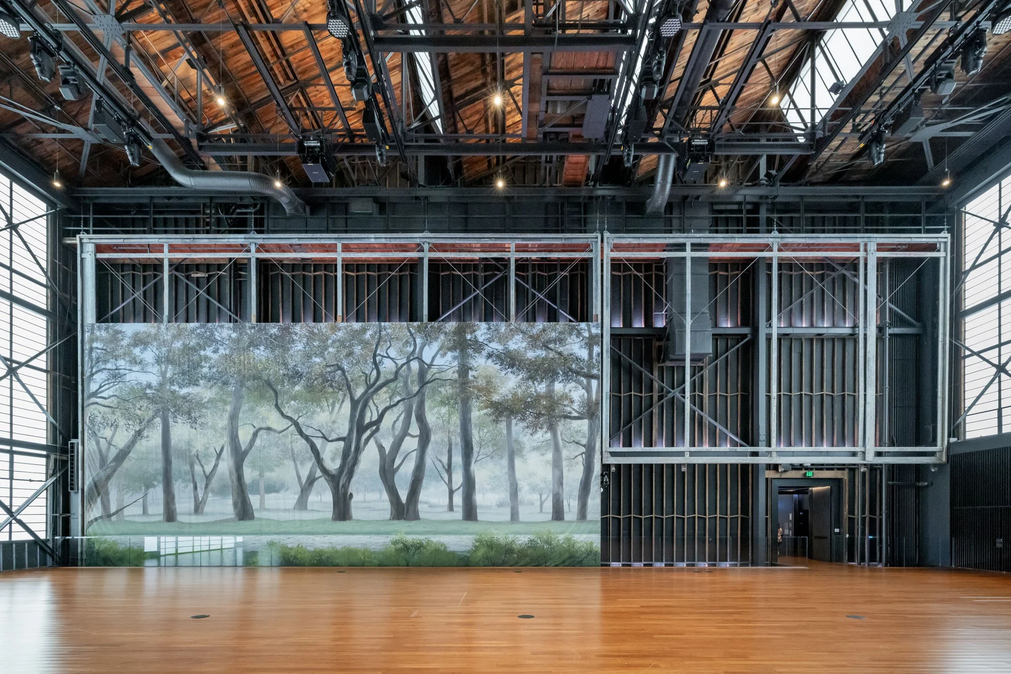 An indoor stage with a large wall mural of trees and a grassy area, steel framing, and a wooden ceiling with exposed beams and lights.