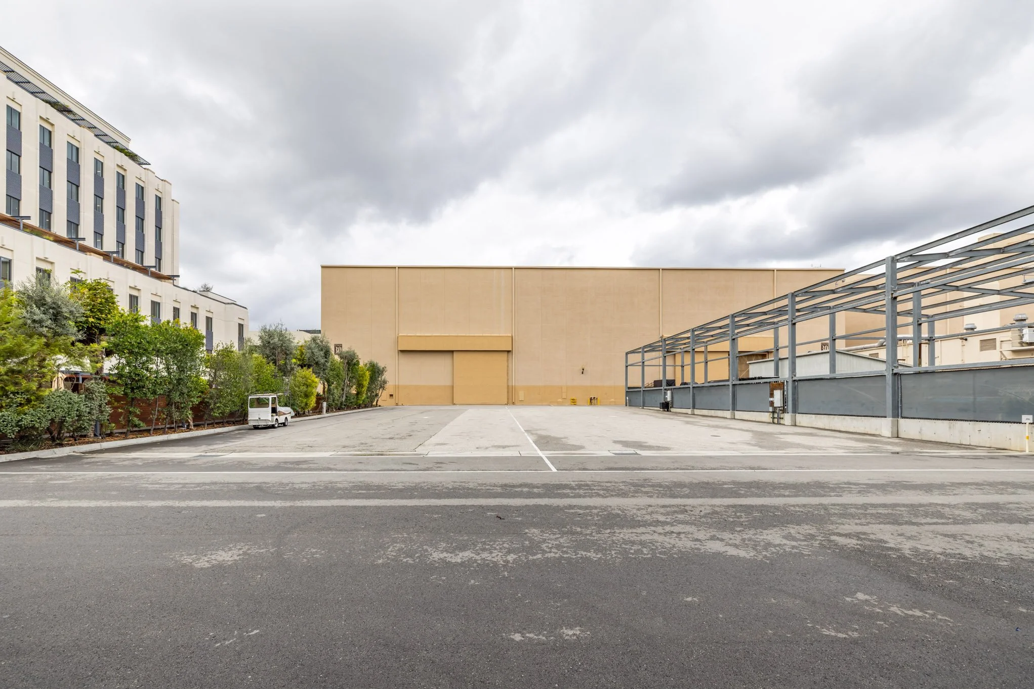 An empty parking lot in an urban area with a beige industrial building and a modern white apartment building in the background, under cloudy skies.