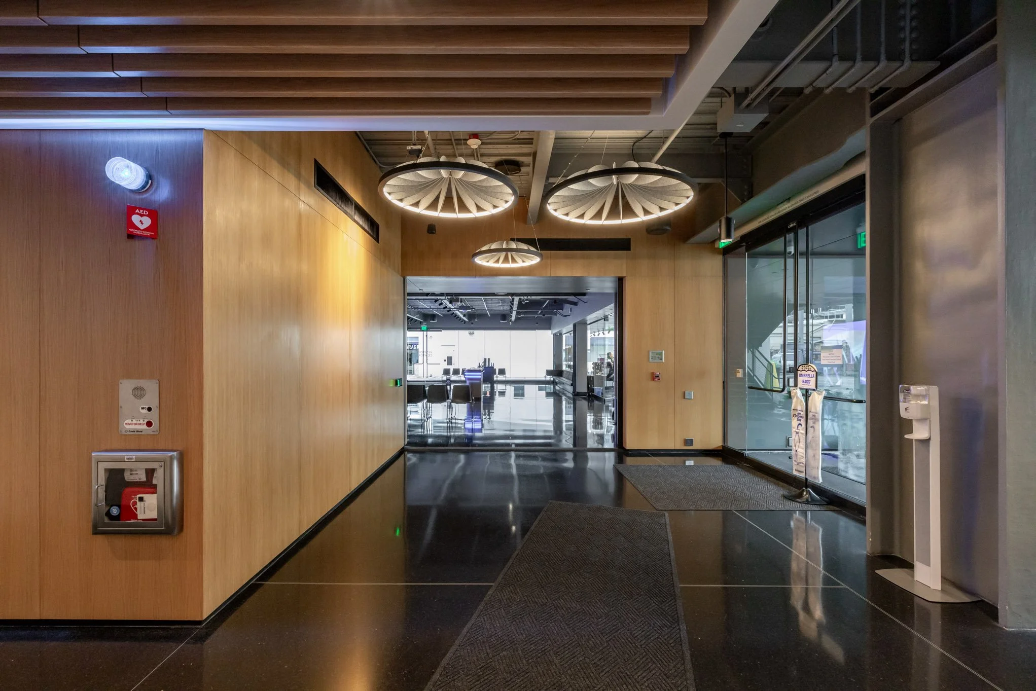 The interior of a modern building with wooden walls, black ceiling, and decorative hanging light fixtures. There are glass doors leading to an outdoor area or another section of the building, with visible seating and lights in the background. Fire sa