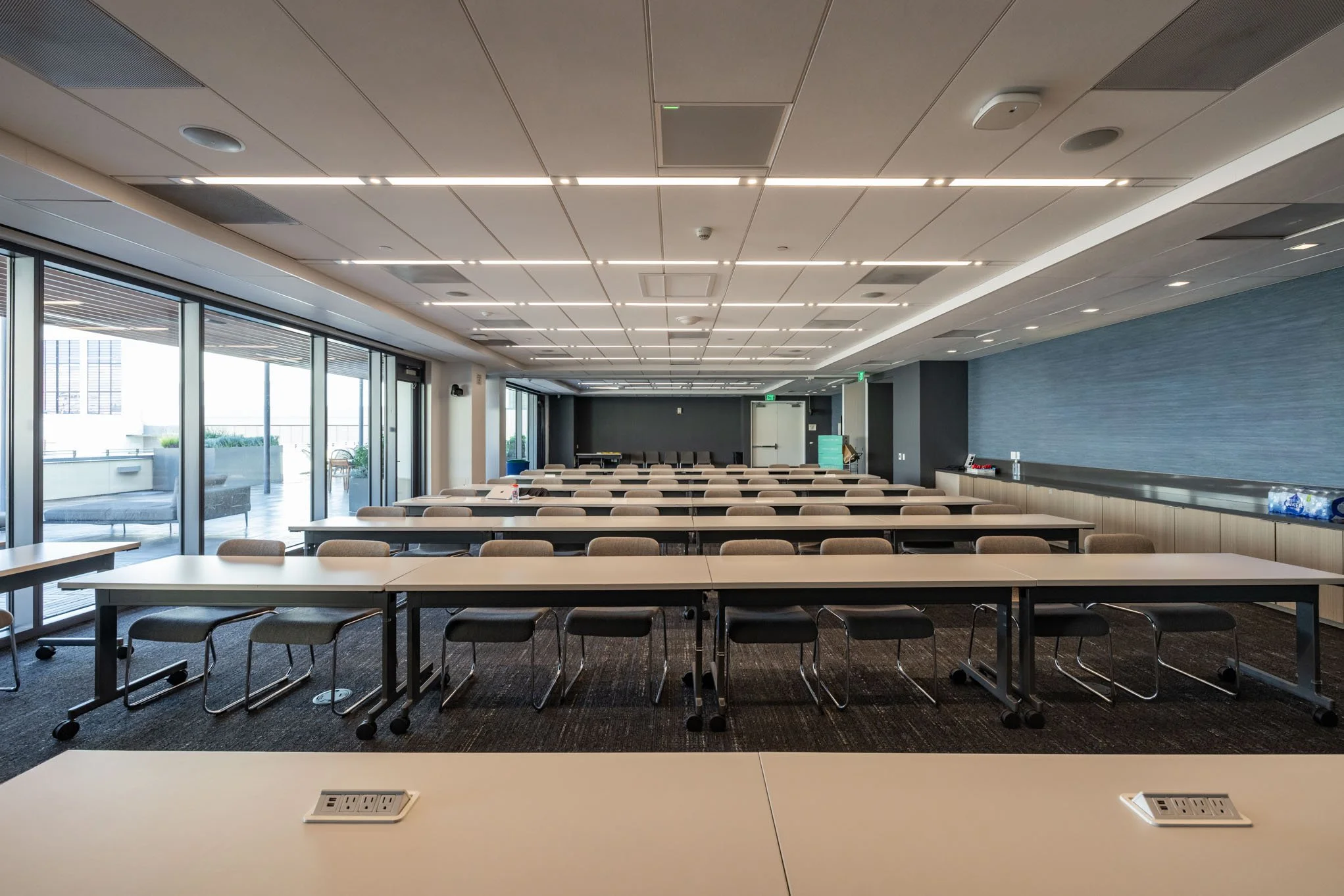 Empty conference room with multiple long tables and chairs, large windows with a balcony outside, and a wall with a textured blue panel.