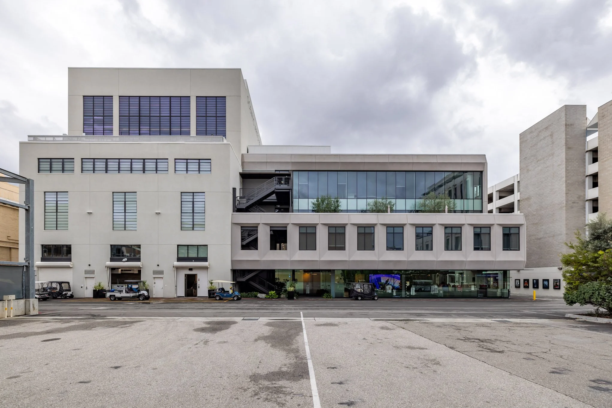 Modern multi-story building with glass windows and a parking lot in front, overcast sky.