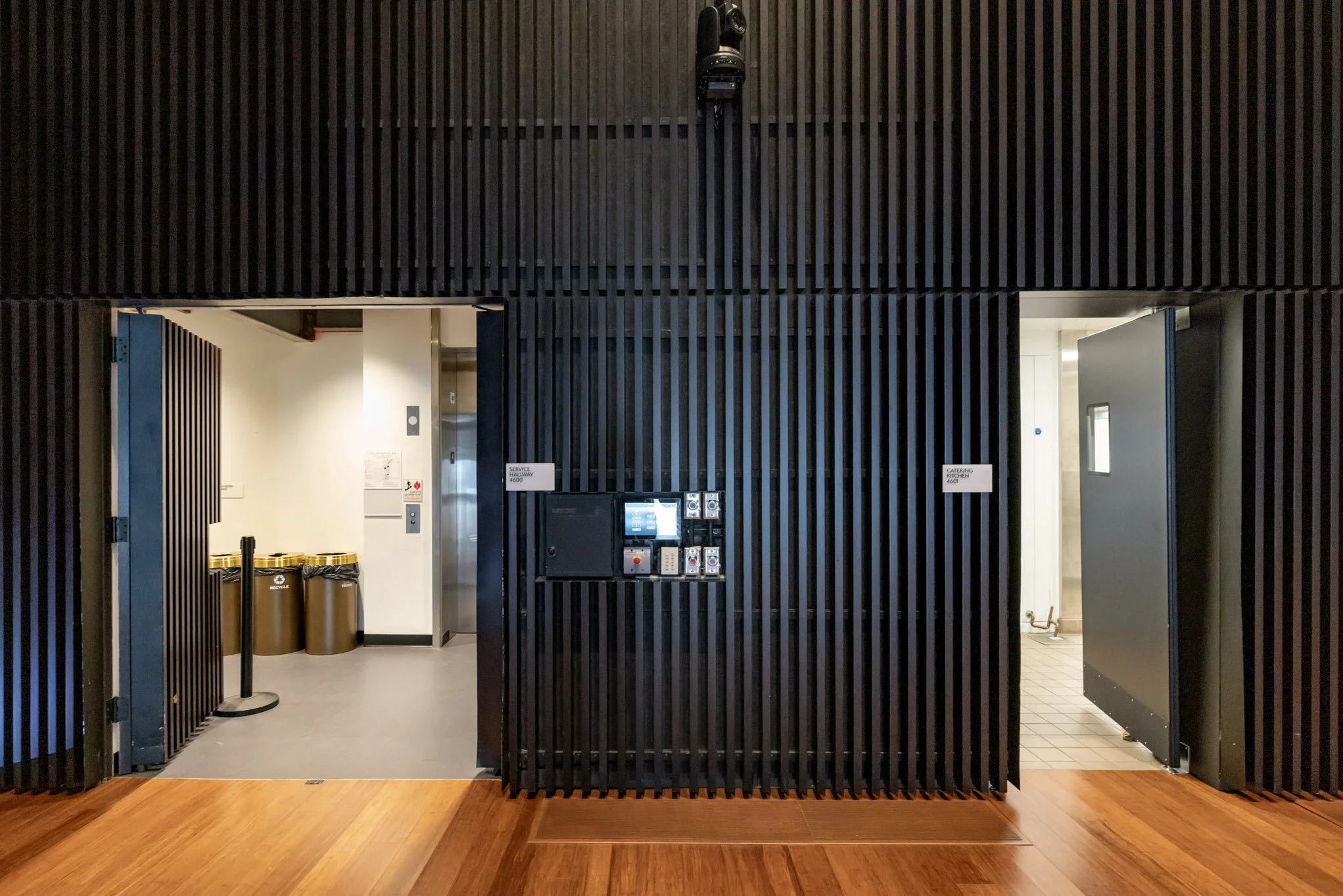 Black wall with two open doors leading to different rooms; room on left has trash bins, room on right has a tiled floor.