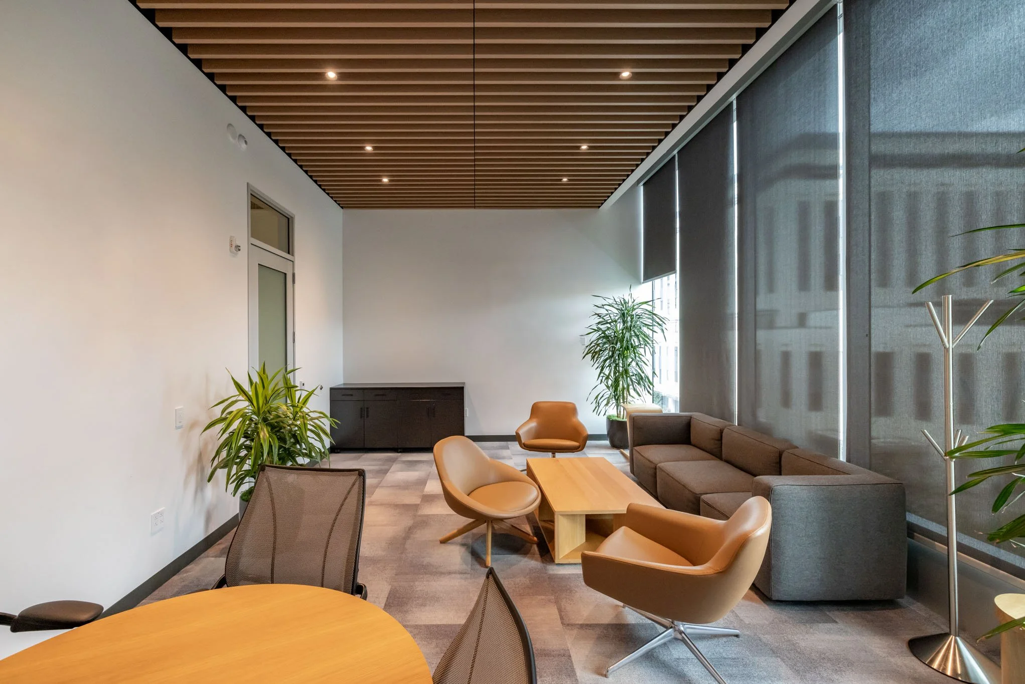Modern office lounge area with a brown sofa, two tan and one beige chairs, a small wooden coffee table, large potted plants, and large windows with dark blinds, wooden slat ceiling, and a gray cabinet in the background.
