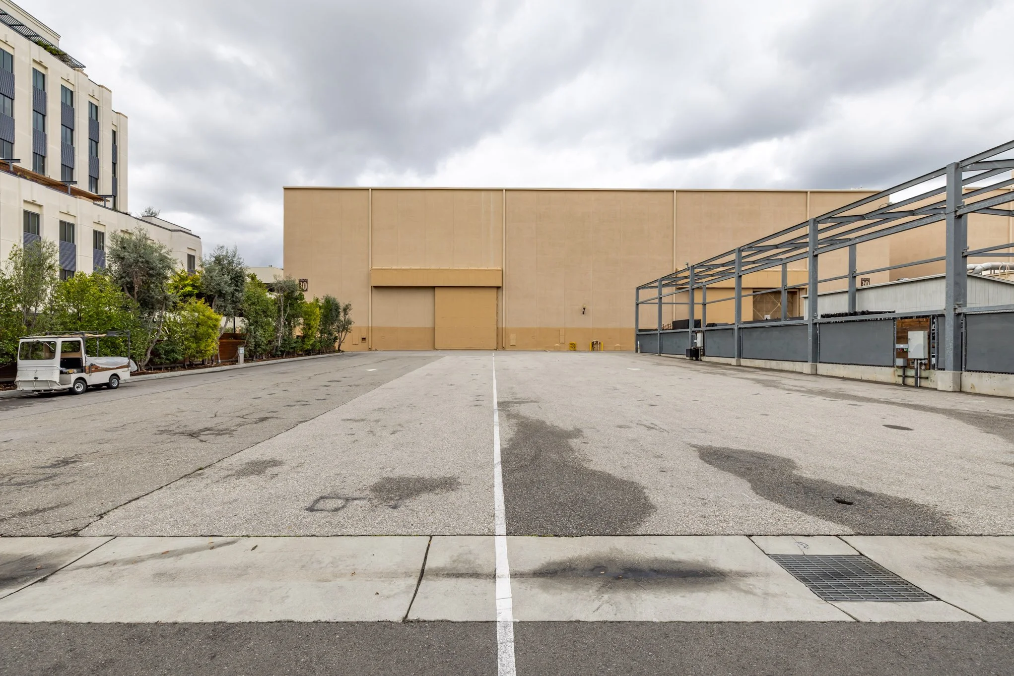 Empty parking lot with a large beige building in the background, cloudy sky overhead, some greenery on the left.