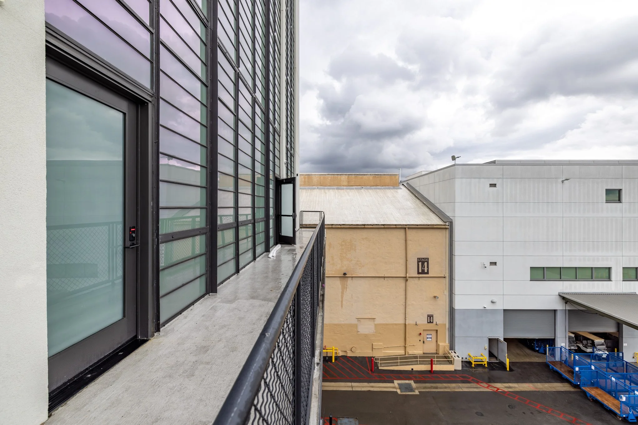 View from a balcony showing glass windows, a door, and a railing on the left, with a loading dock, warehouse building, and cloudy sky in the background.