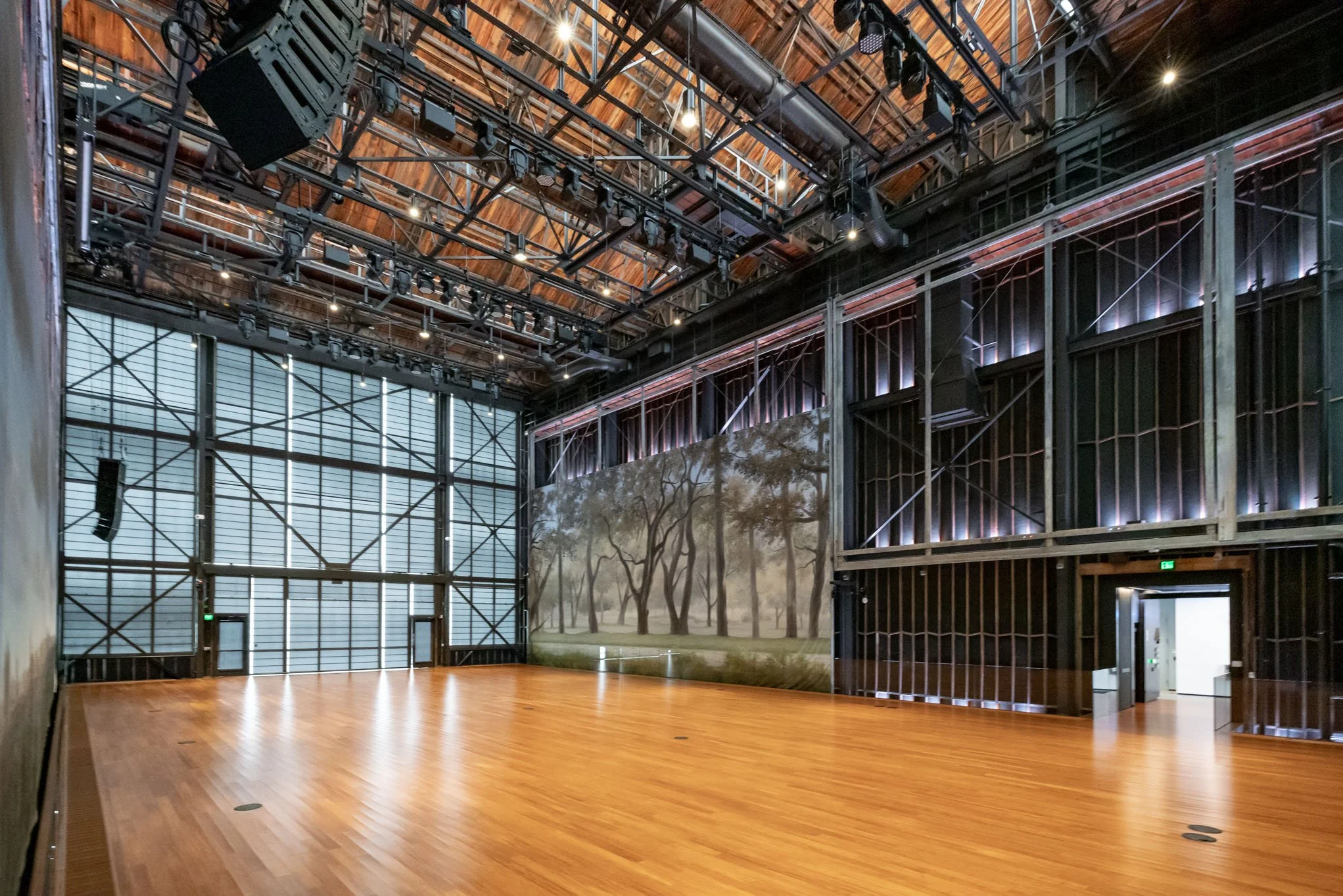 Empty indoor stage with wooden floor, large wall mural of trees, and visible stage lighting and rigging overhead.