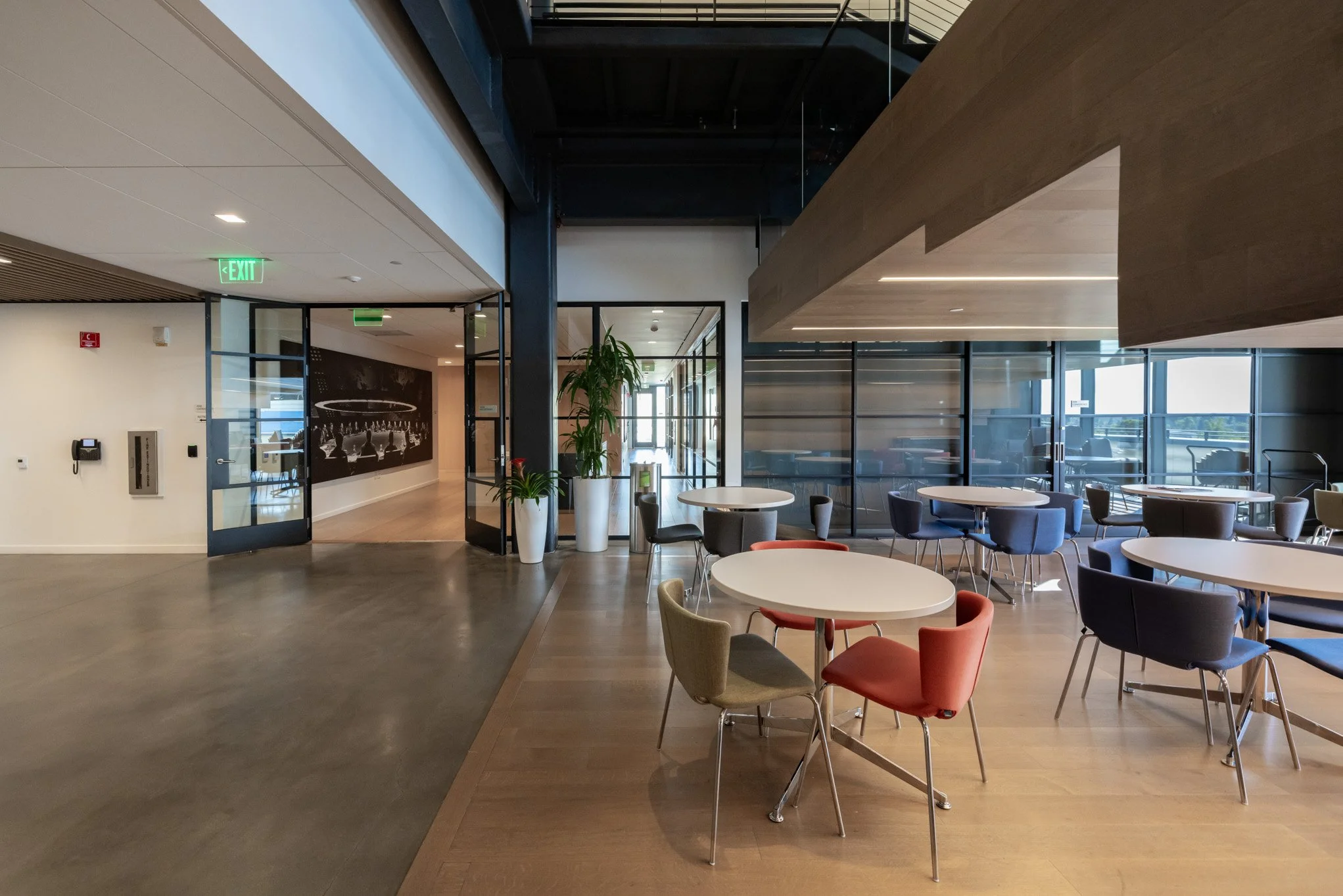 An empty modern lounge area with round tables and colorful chairs, large windows, potted plants, and exit signs.