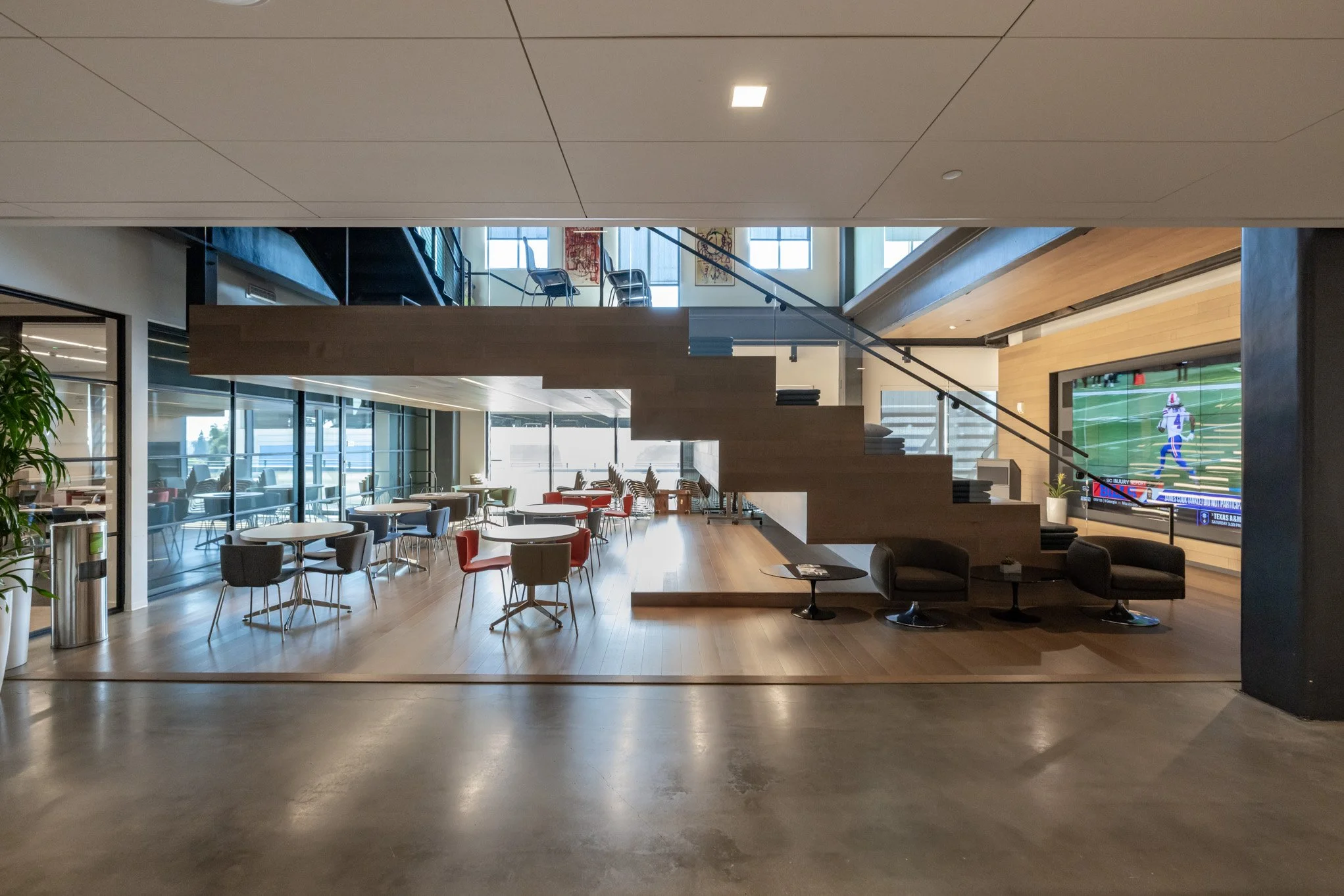 Interior of a modern, open-concept lounge or common area with multiple tables and chairs, a staircase leading to an upper level, large windows allowing natural light, a large flat-screen TV showing a football game, and contemporary decor.