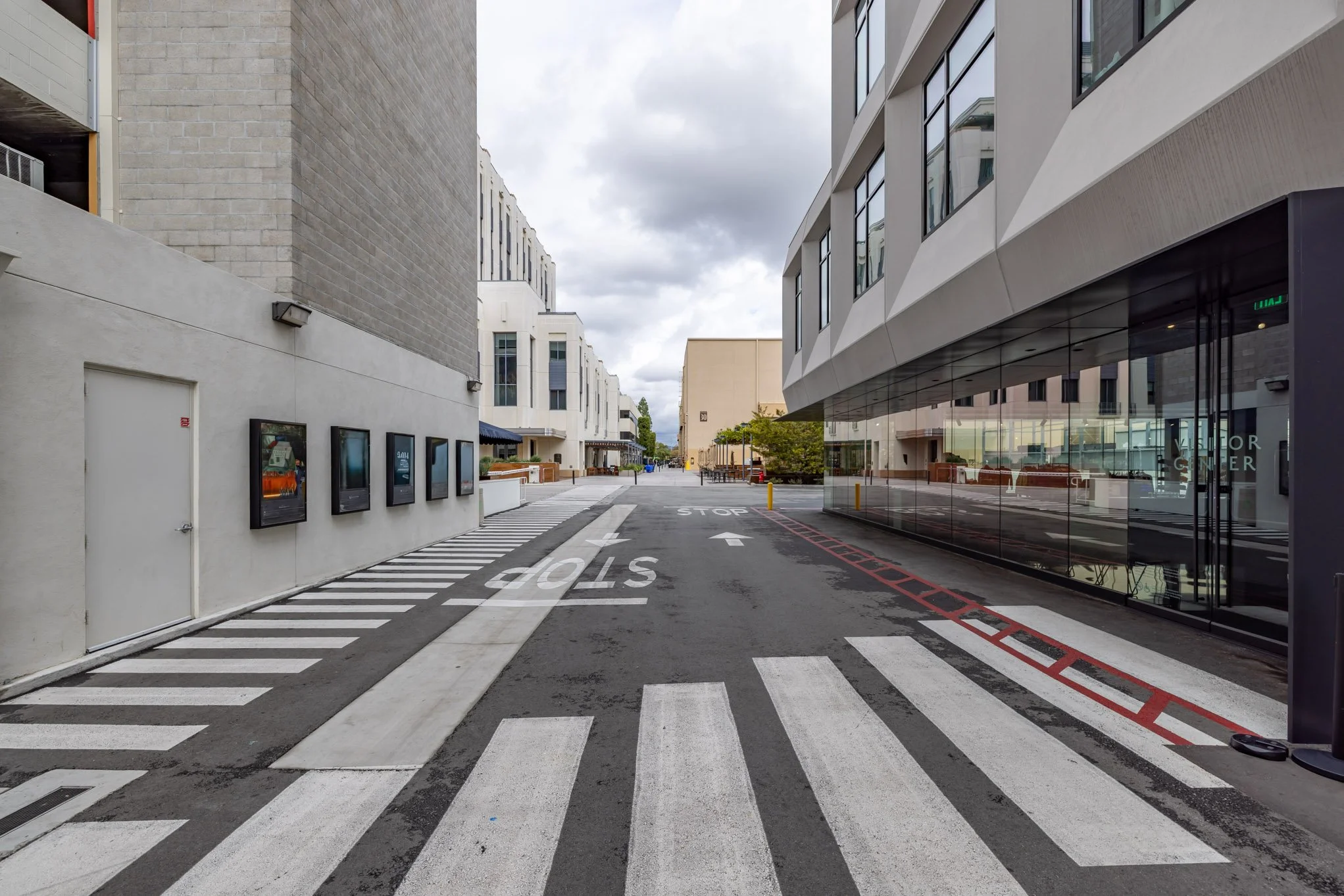 Empty city street with buildings on both sides, crosswalks, stop signs, and reflections on glass windows; cloudy sky overhead.