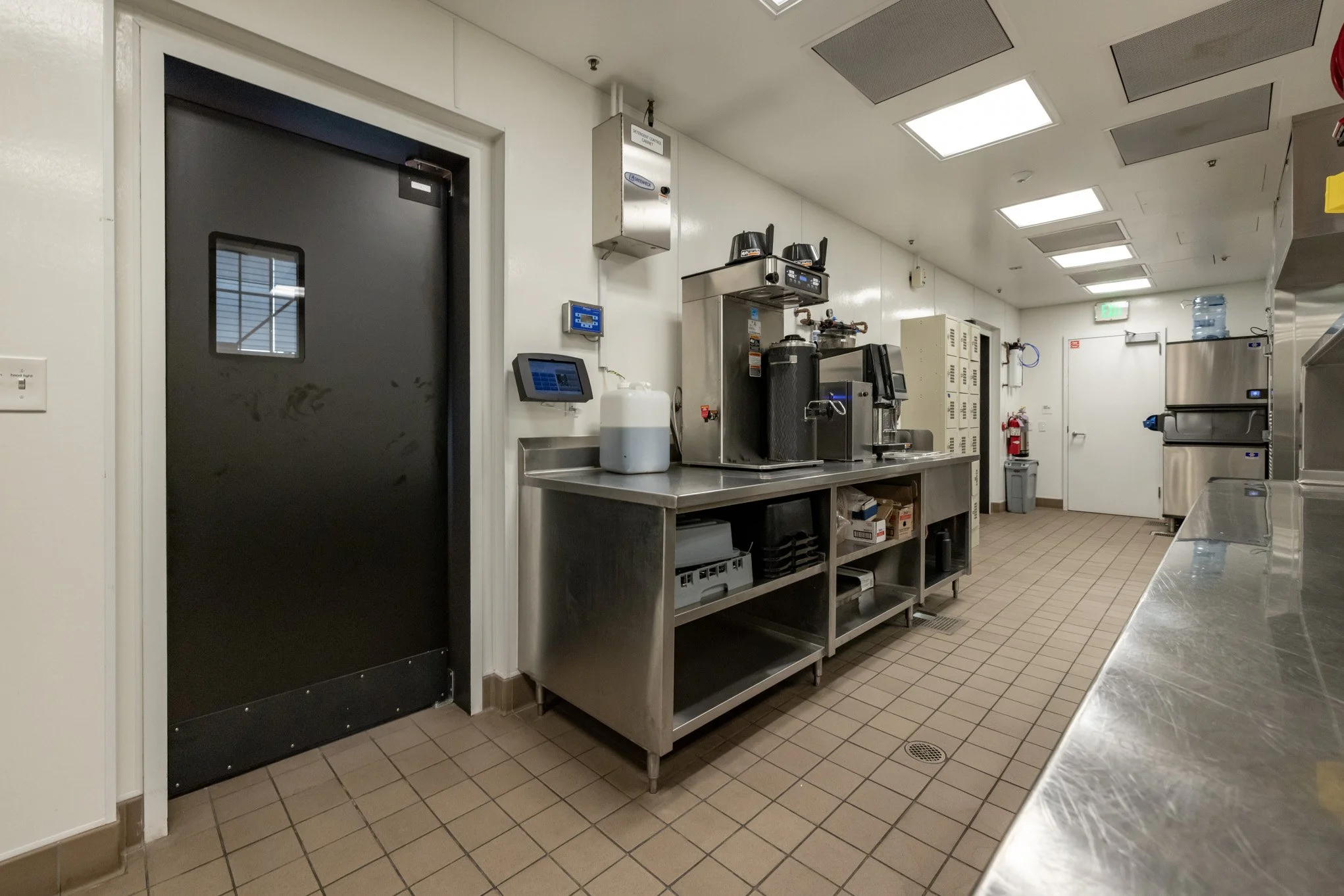 Commercial kitchen with stainless steel counters and appliances, fire extinguisher, water cooler, and a black door.
