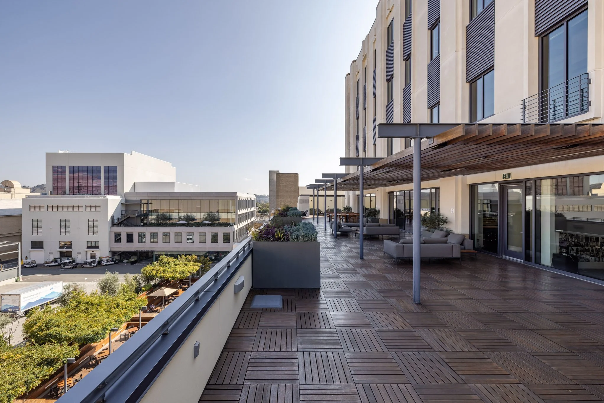 A spacious rooftop balcony in an urban setting with wooden flooring, modern outdoor furniture, potted plants, and a view of surrounding buildings and trees under a clear sky.