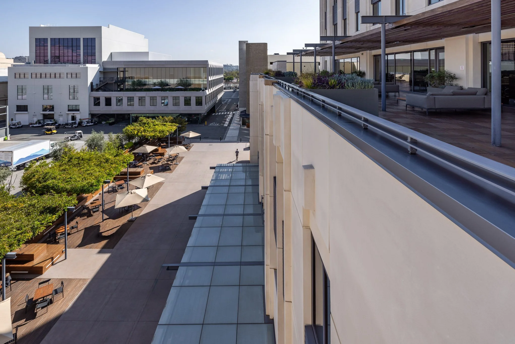 Top view of an outdoor urban space with seating areas, trees, and buildings, showing a modern balcony with patio furniture and planters.