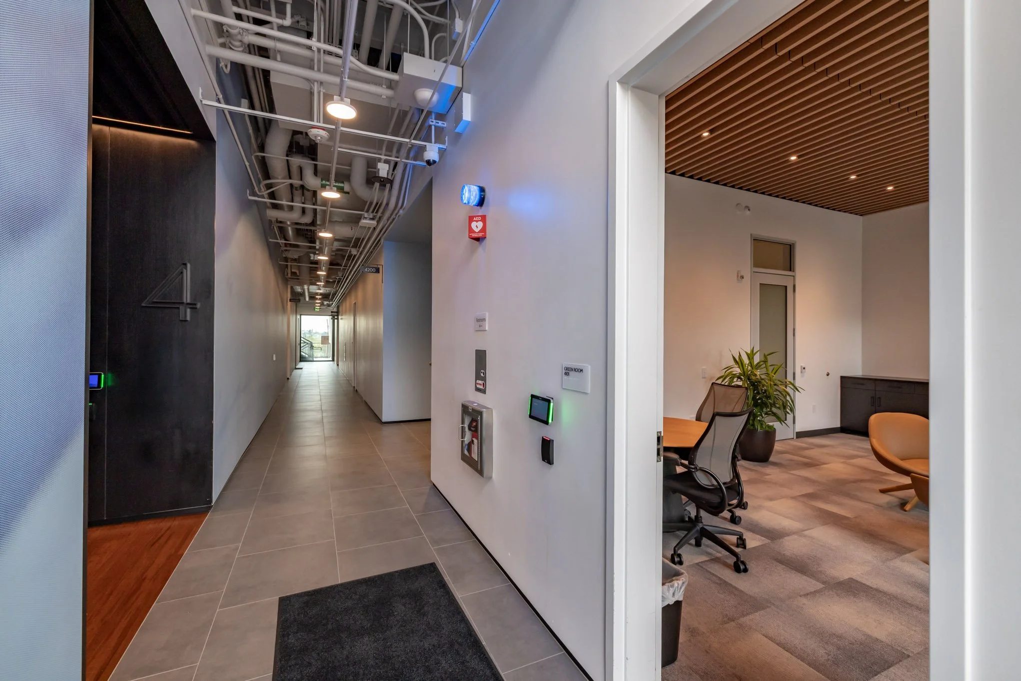 Interior view of a modern building with a hallway on the left side featuring exposed ceiling pipes and a doorway on the right leading to a room with chairs and a table.