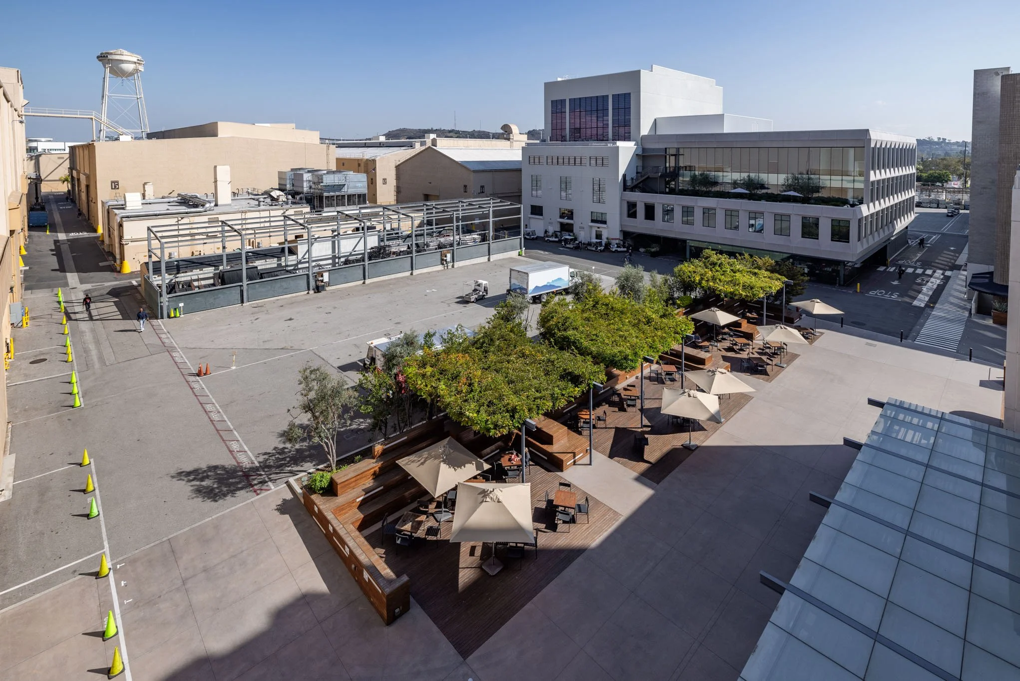 Empty outdoor seating area with umbrellas and trees in an urban plaza, surrounded by modern buildings.